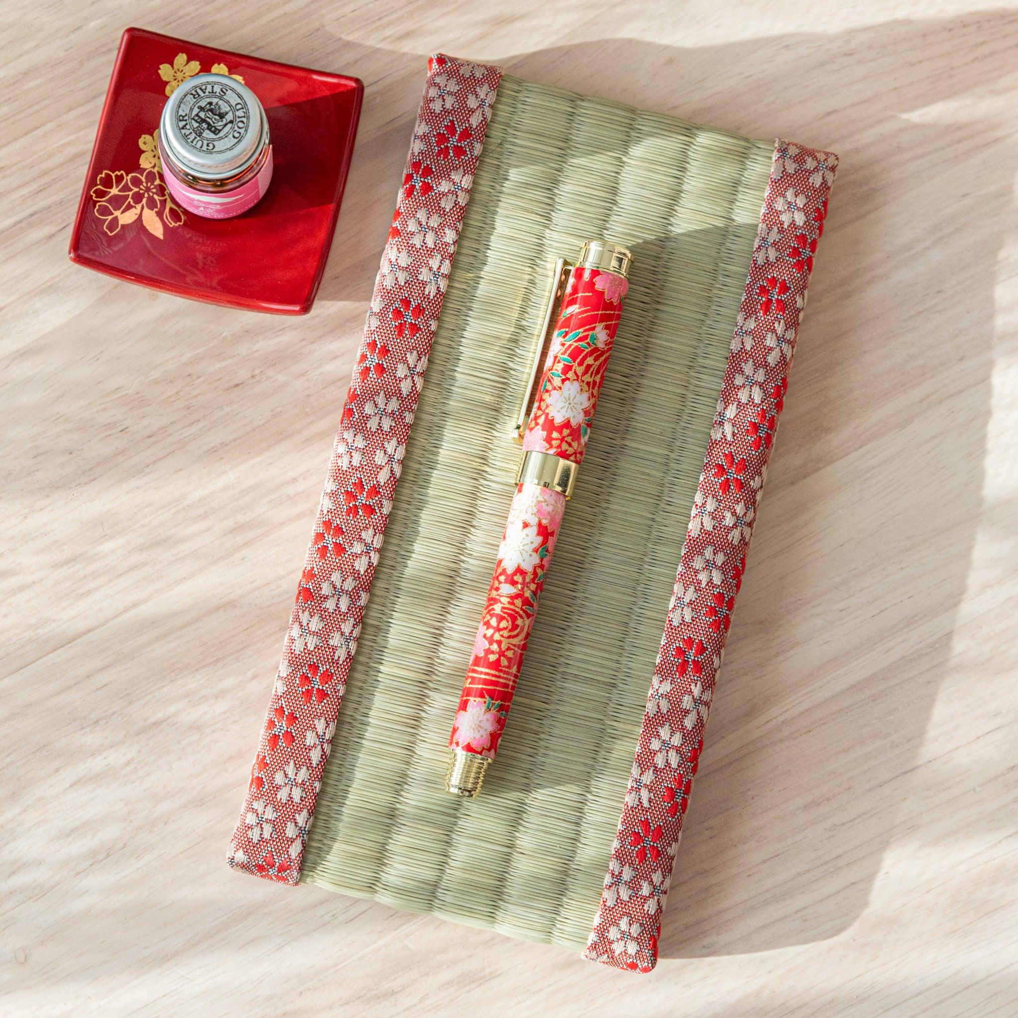 A red floral Japanese stationery fountain pen placed on a tatami-style mat with a red dish and ink bottle in the background.  