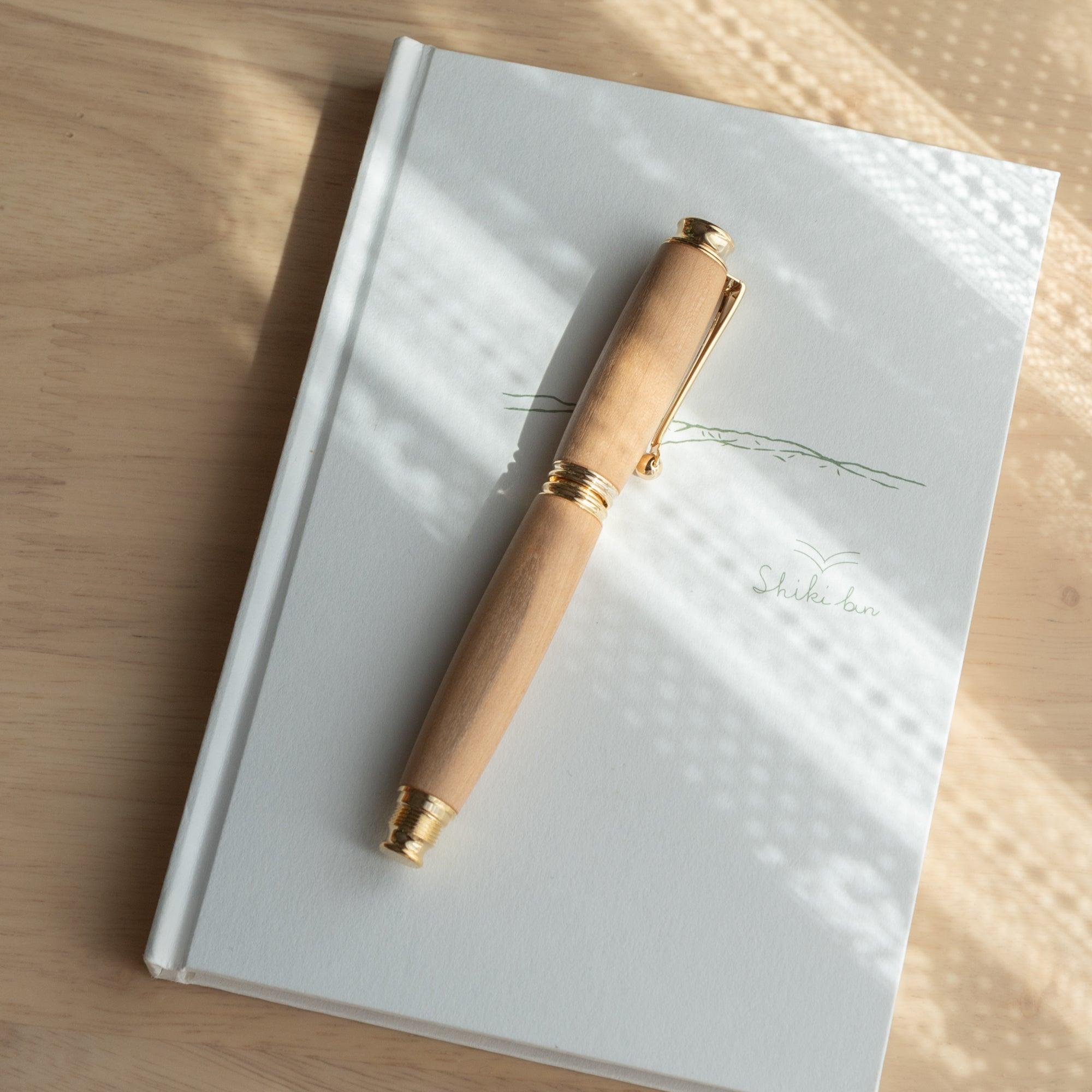 A Japanese fountain pen made of wood with gold accents resting horizontally on a white notebook under soft sunlight, highlighting its elegant design.