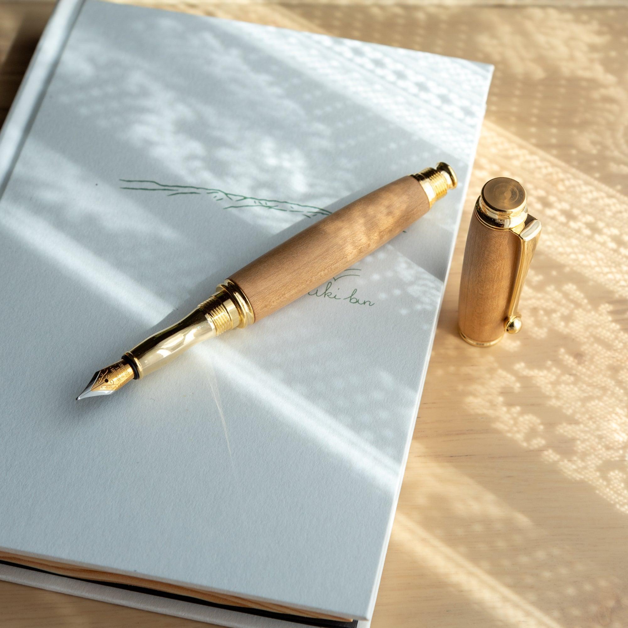 A Japanese fountain pen with a gold nib, wooden barrel, and detached cap placed on a white notebook, surrounded by intricate light and shadow patterns.