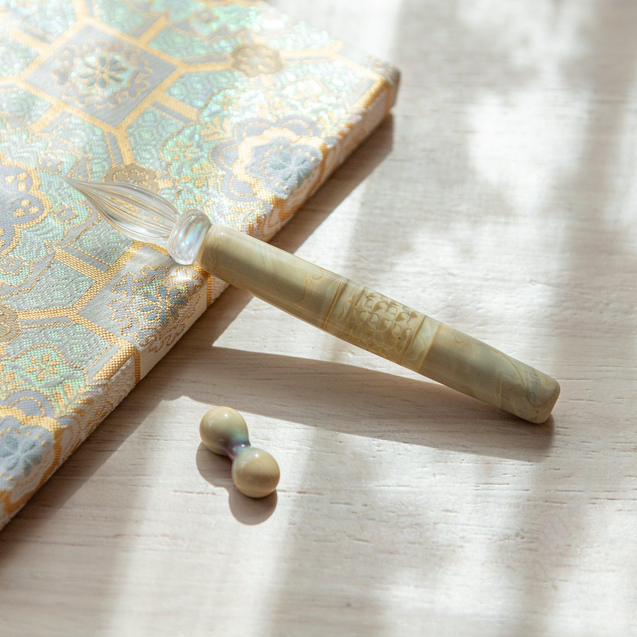 Ivory glass pen with a shippo design, paired with a matching glass pen rest. Displayed on a wooden surface beside a Nishijin textile notebook.