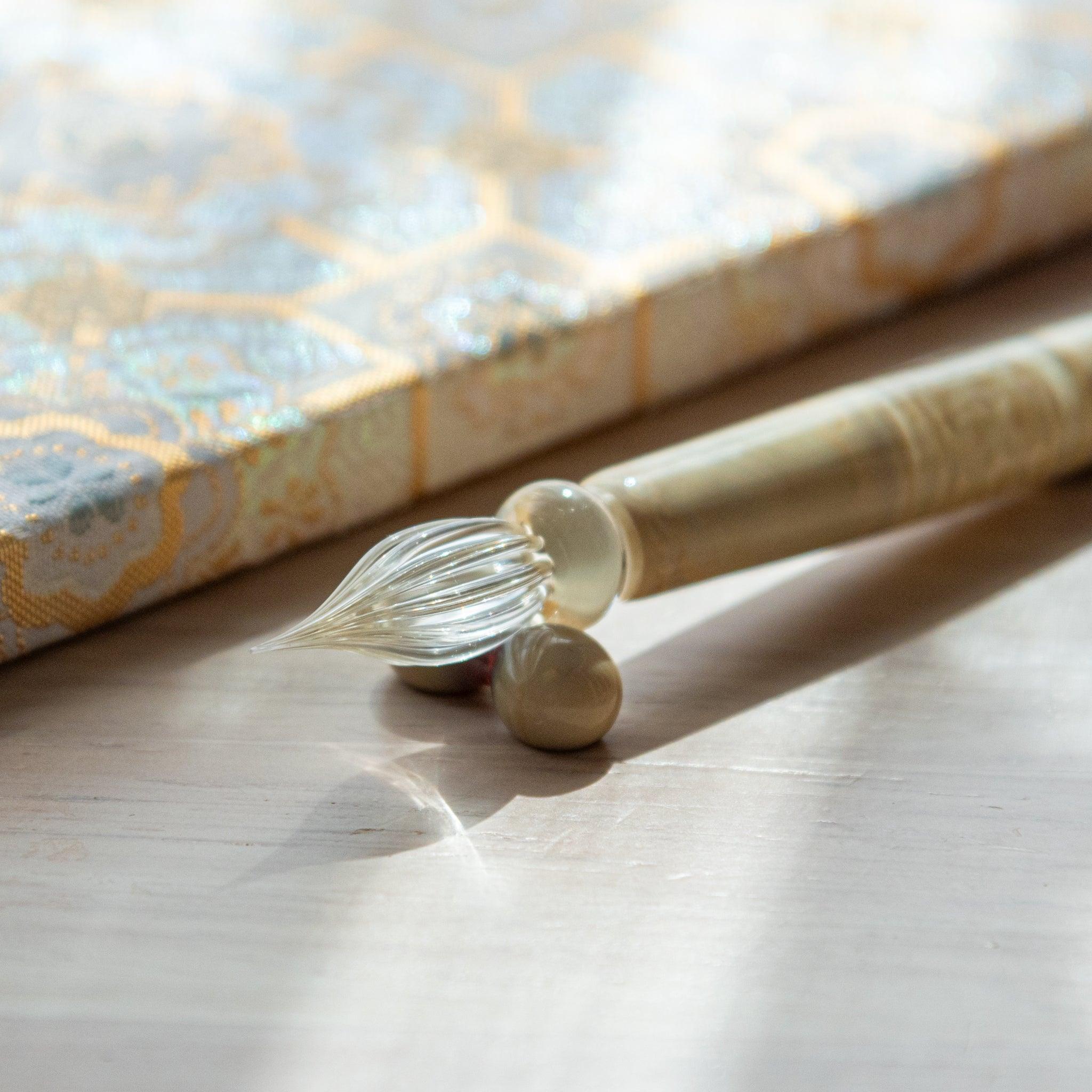 Ivory glass pen with a shippo design, paired with a matching glass pen rest. Displayed on a wooden surface beside a Nishijin textile notebook.