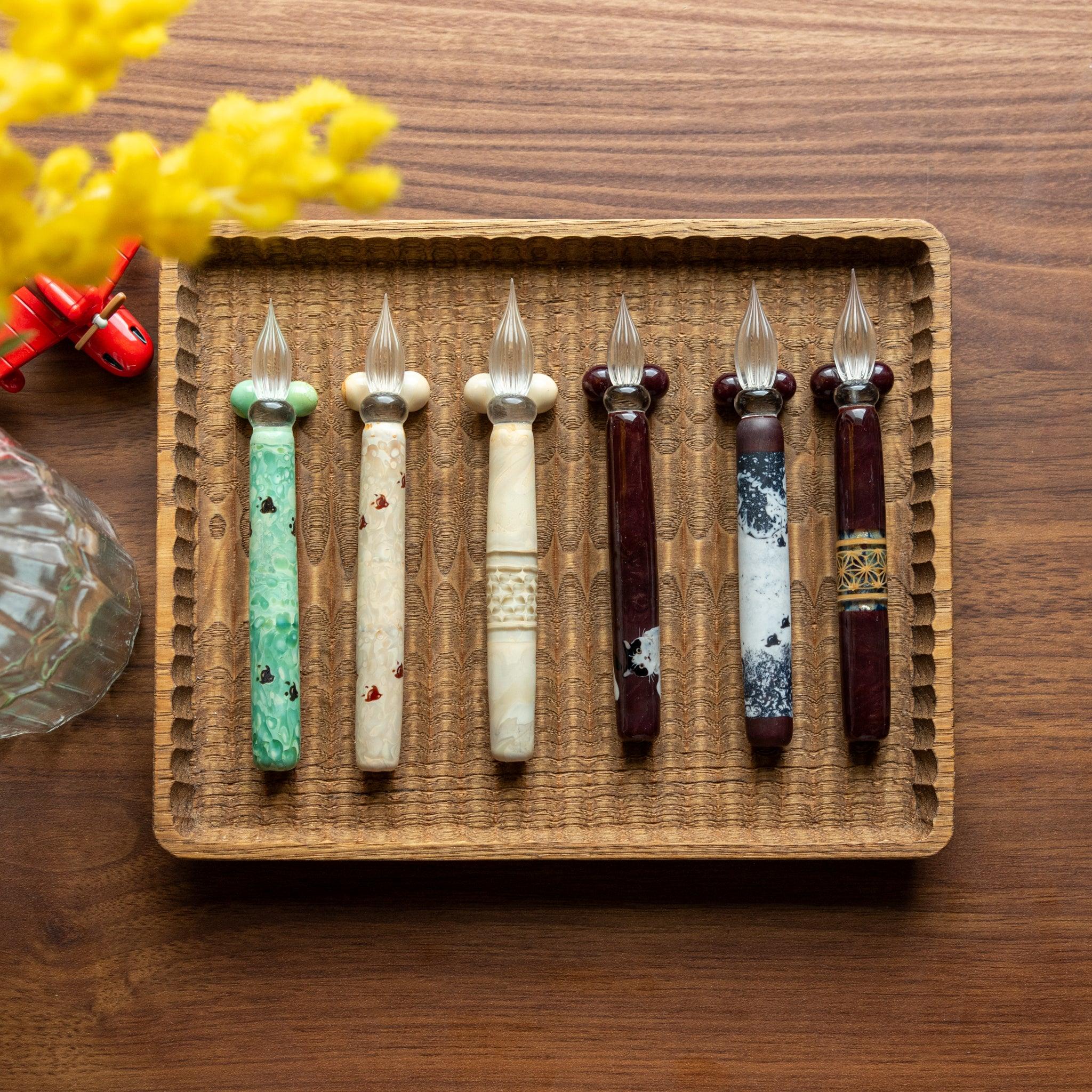 Six glass pens with traditional designs displayed on a hand-carved wooden pen tray. Each pen showcases unique craftsmanship.