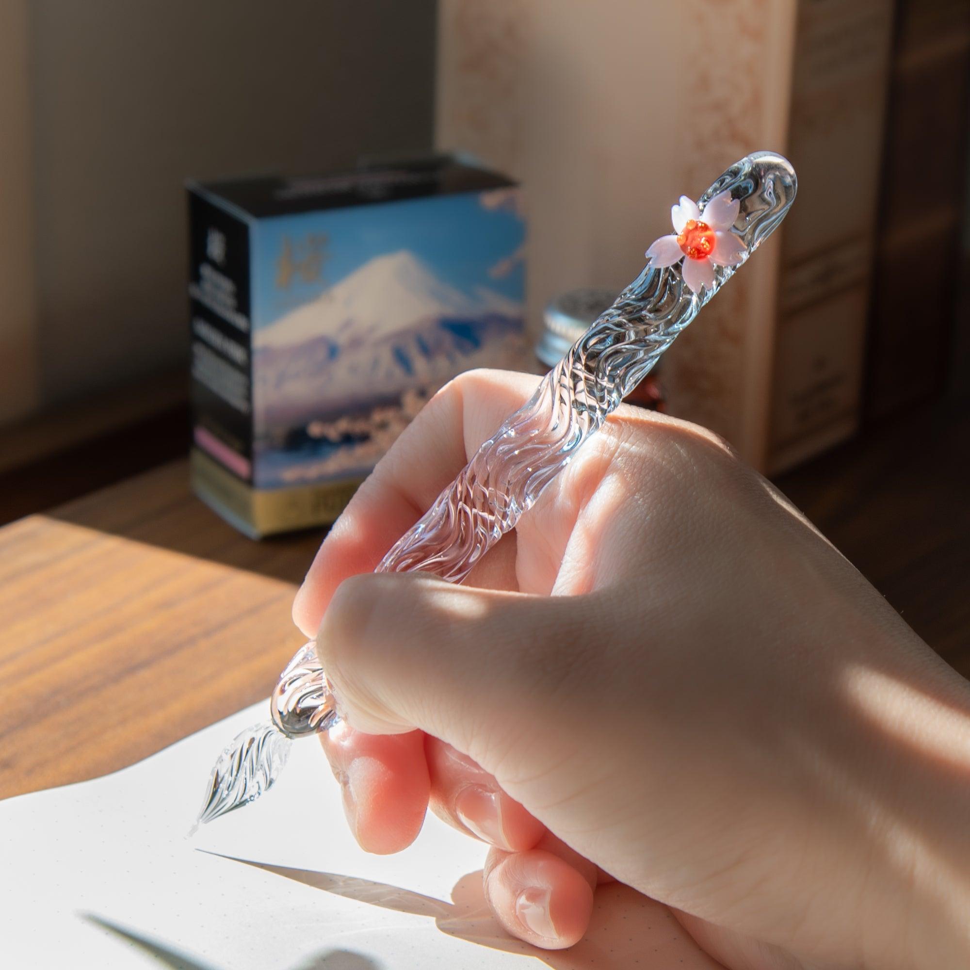 The sakura motif glass pen is held in hand, with light streaming in and a box with Mount Fuji art in the background.