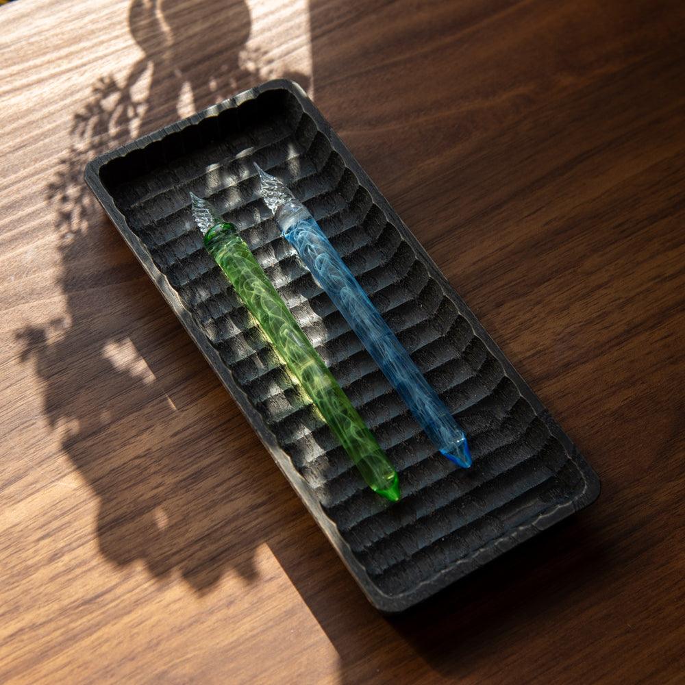 Two glass pens, one green and one blue, resting in a textured tray on a wooden surface. Shadows of nearby leaves are cast over the scene.