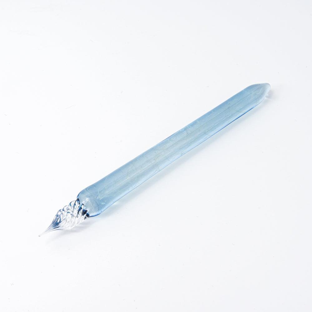 A light blue glass pen with delicate smoke patterns, lying on a white surface, showcasing its transparent nib and sleek look.