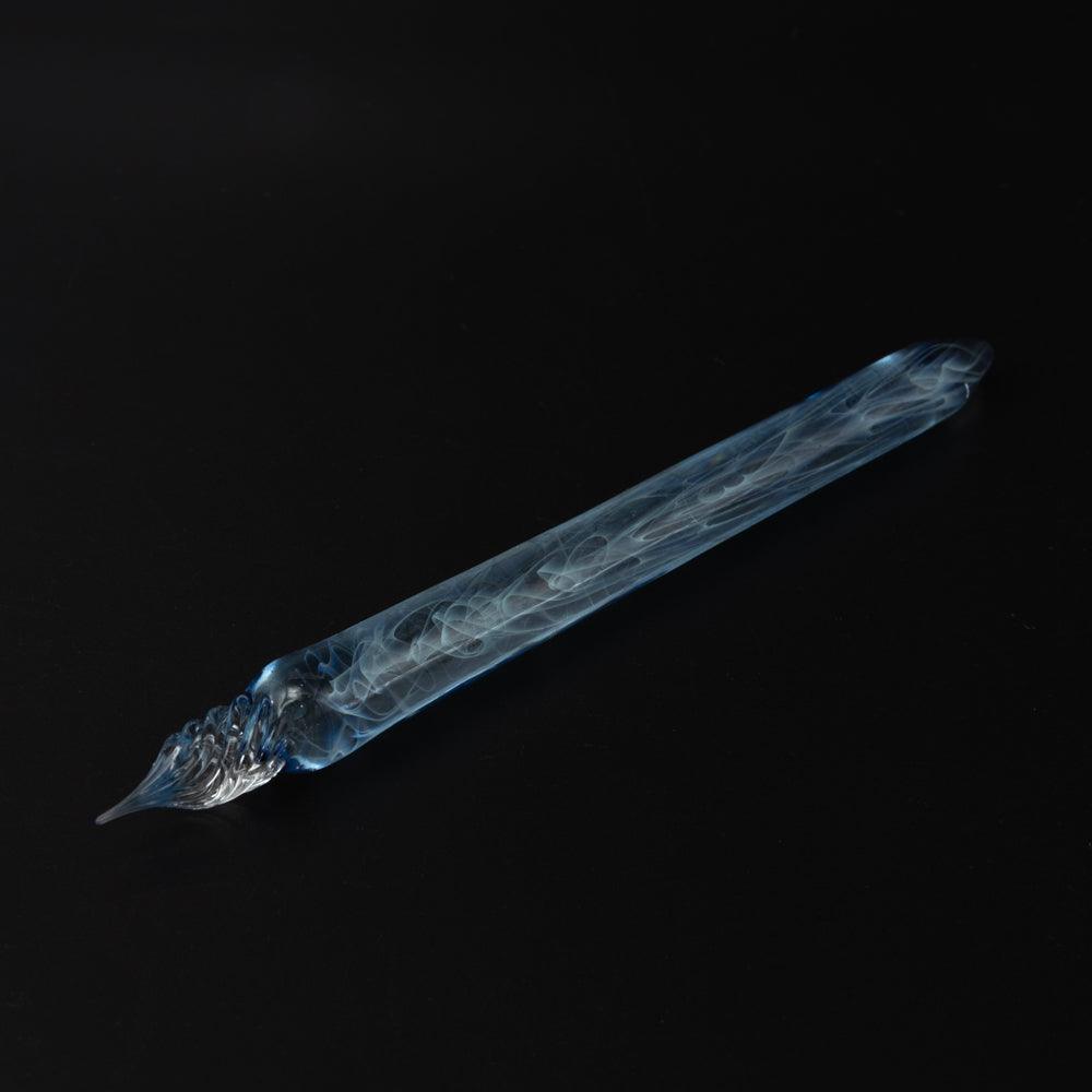 A blue glass pen with smoke swirls on a black background. The clear glass nib and blue hues highlight the pen's elegant design.