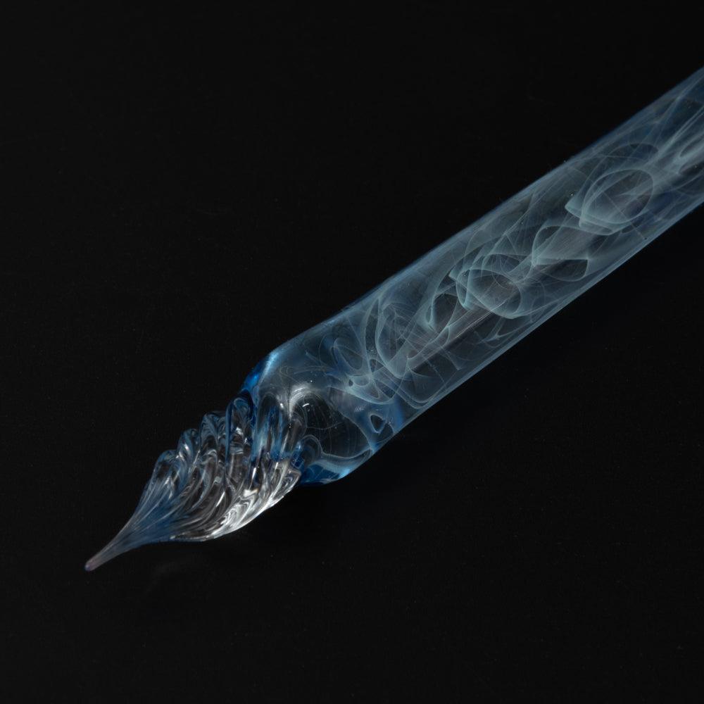 A blue glass pen with smoke swirls on a black background. The clear glass nib and blue hues highlight the pen's elegant design.