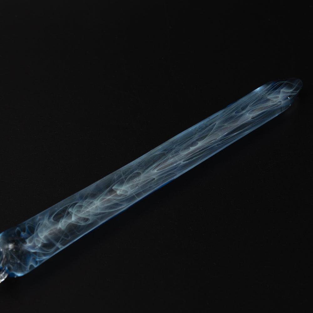 A blue glass pen with smoke swirls on a black background. The clear glass nib and blue hues highlight the pen's elegant design.