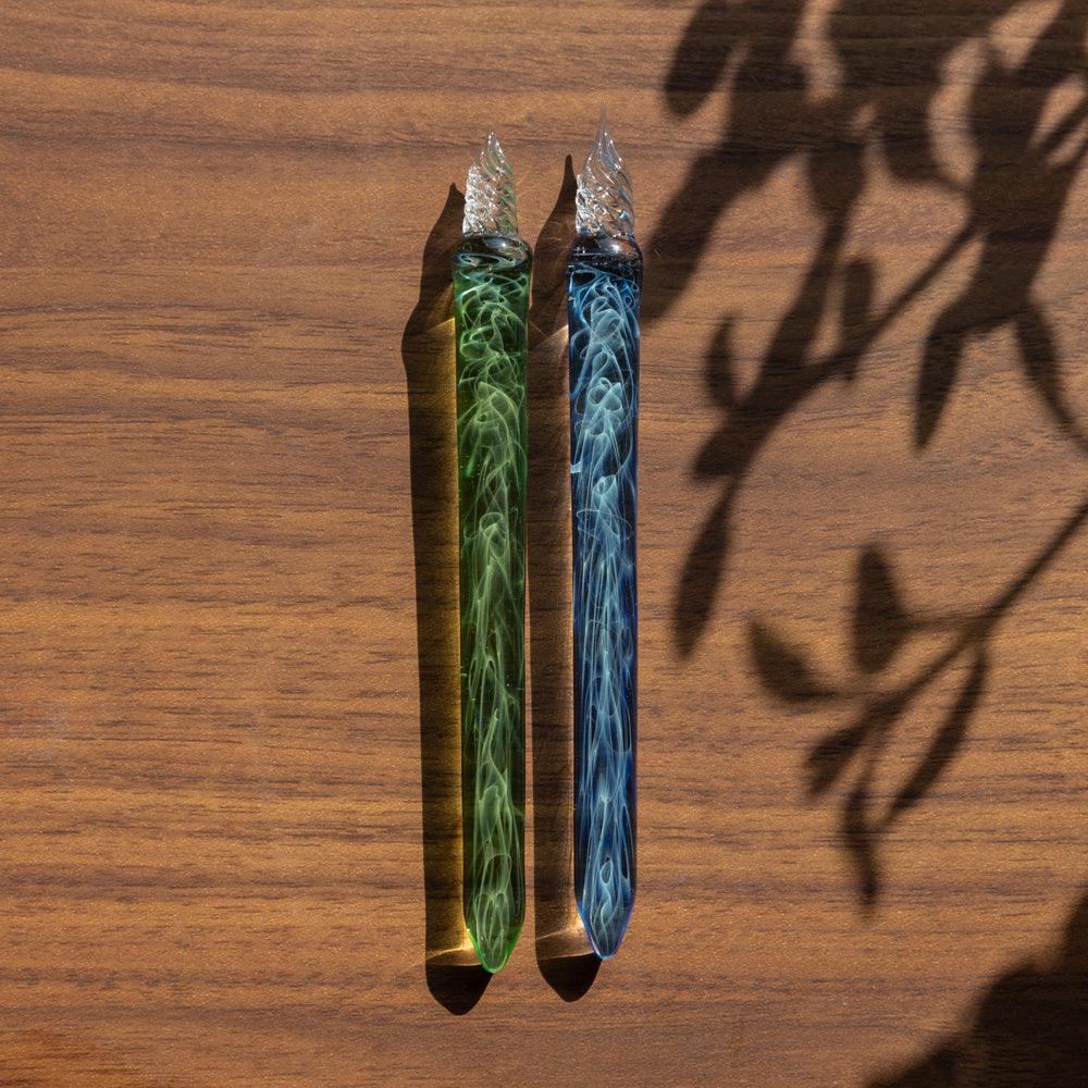 Two glass pens with green and blue swirling designs, lying side by side on a wooden surface with shadows from nearby leaves.