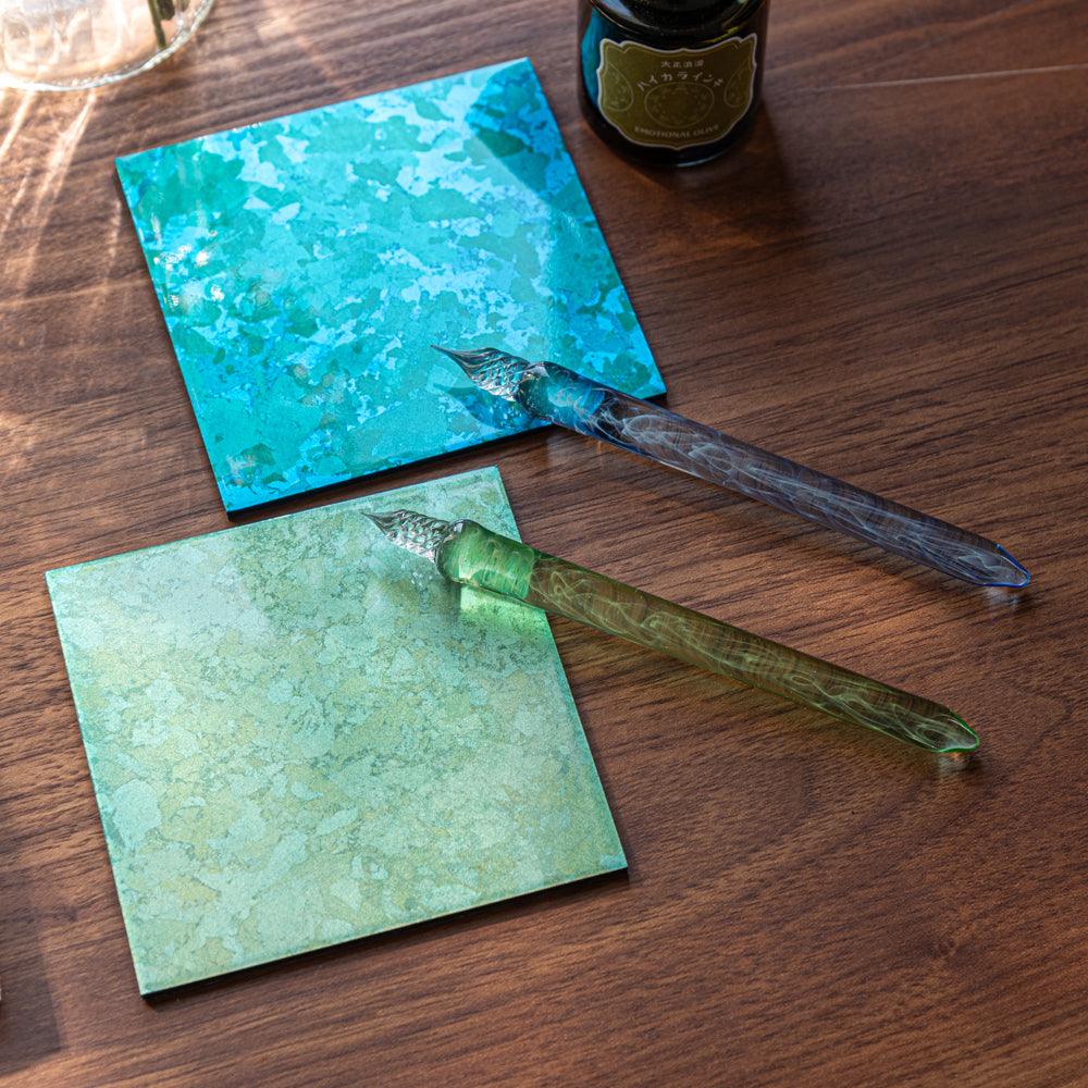 Two glass pens, green and blue, resting on colorful square tiles, with an ink bottle nearby. The tiles add a vibrant background.