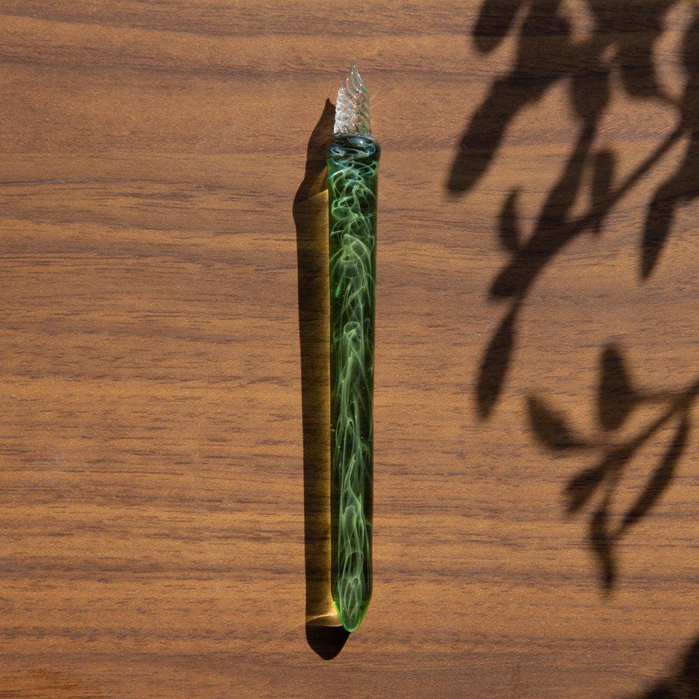 Green glass pen with swirling designs resting on a wooden surface, casting soft shadows from nearby foliage under sunlight.