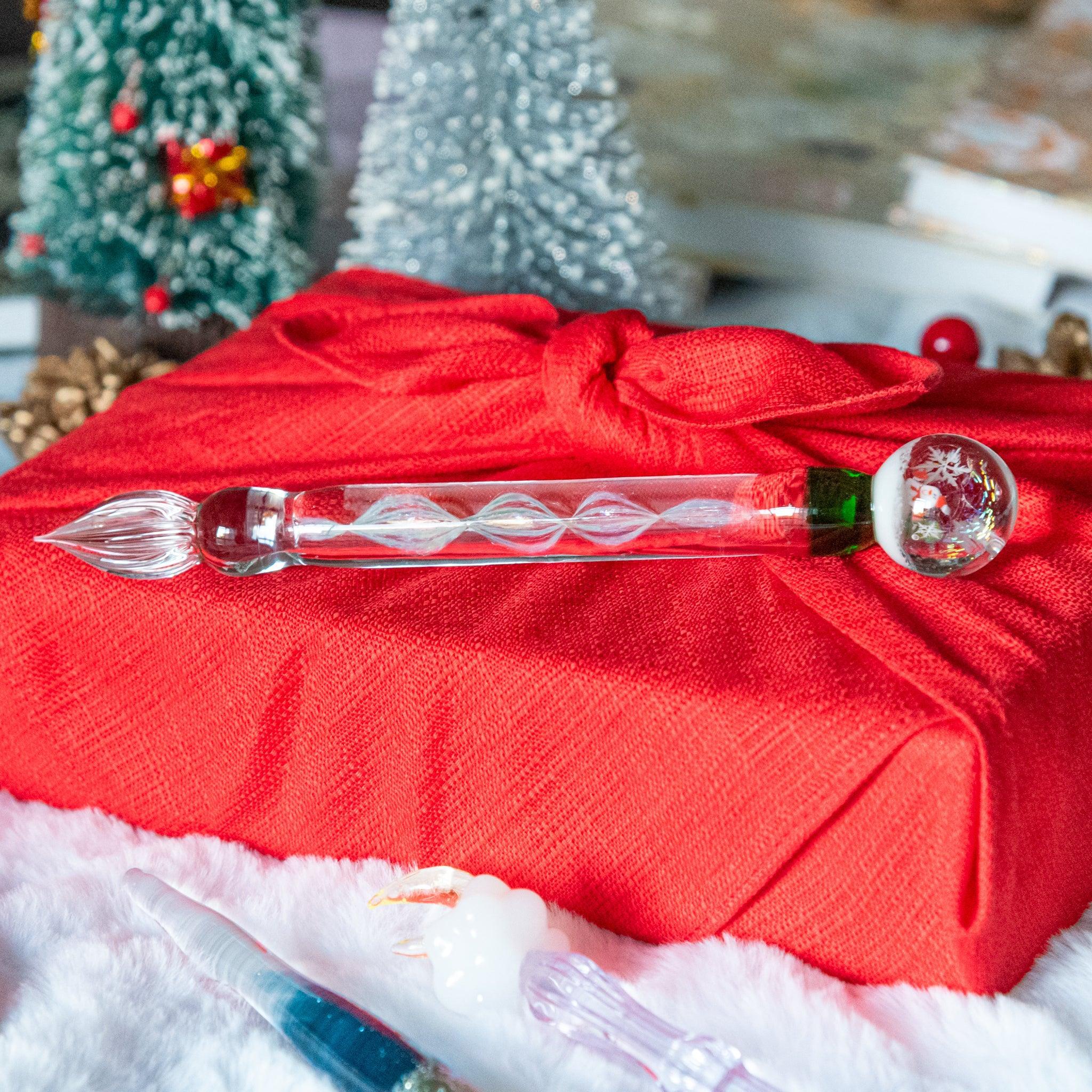 A glass pen with a snow globe-like design featuring a snowman inside rests on a red cloth-wrapped gift, surrounded by festive decorations.