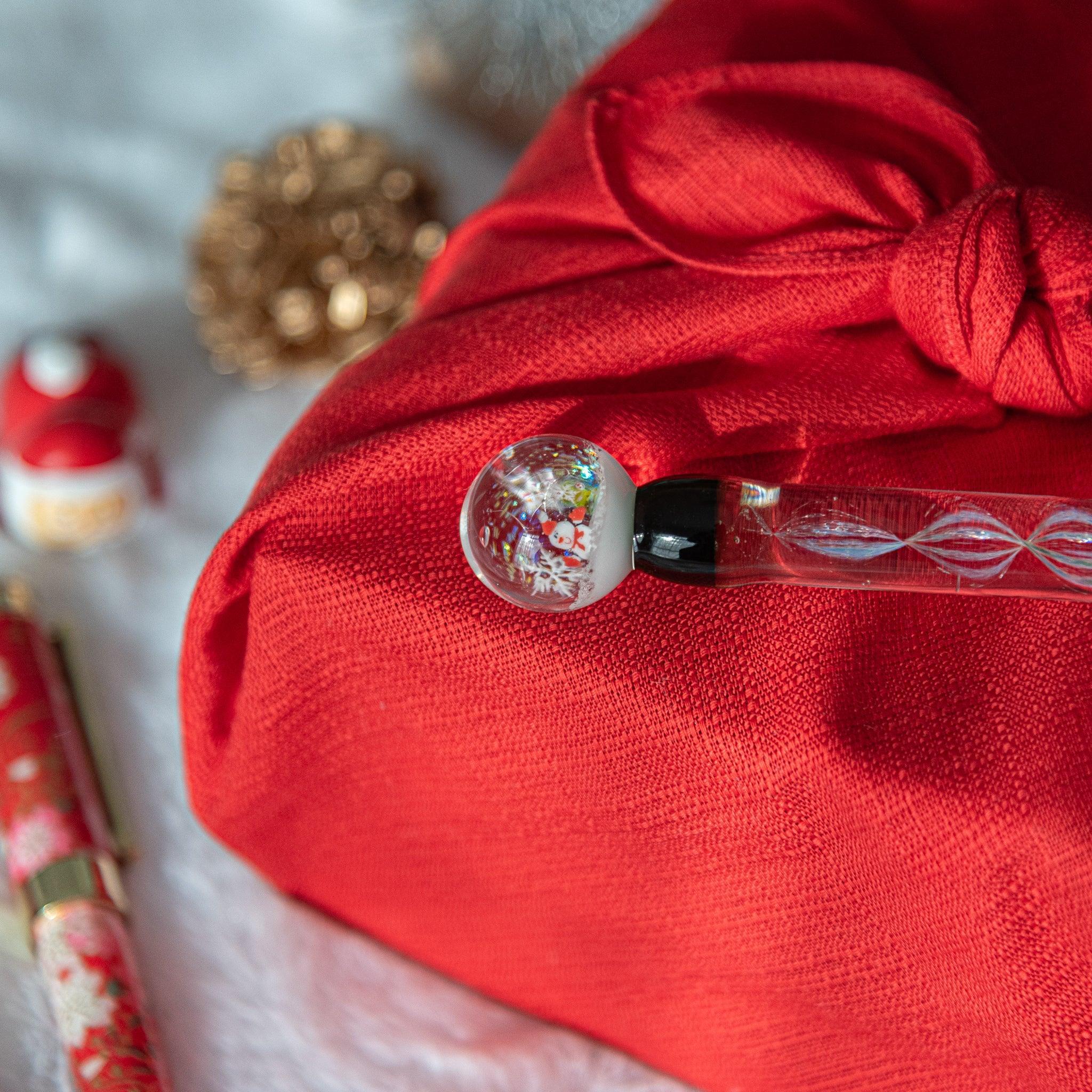 A glass pen with a snow globe-inspired cap containing a snowman lies on a red cloth-wrapped box, accompanied by holiday-themed ornaments.