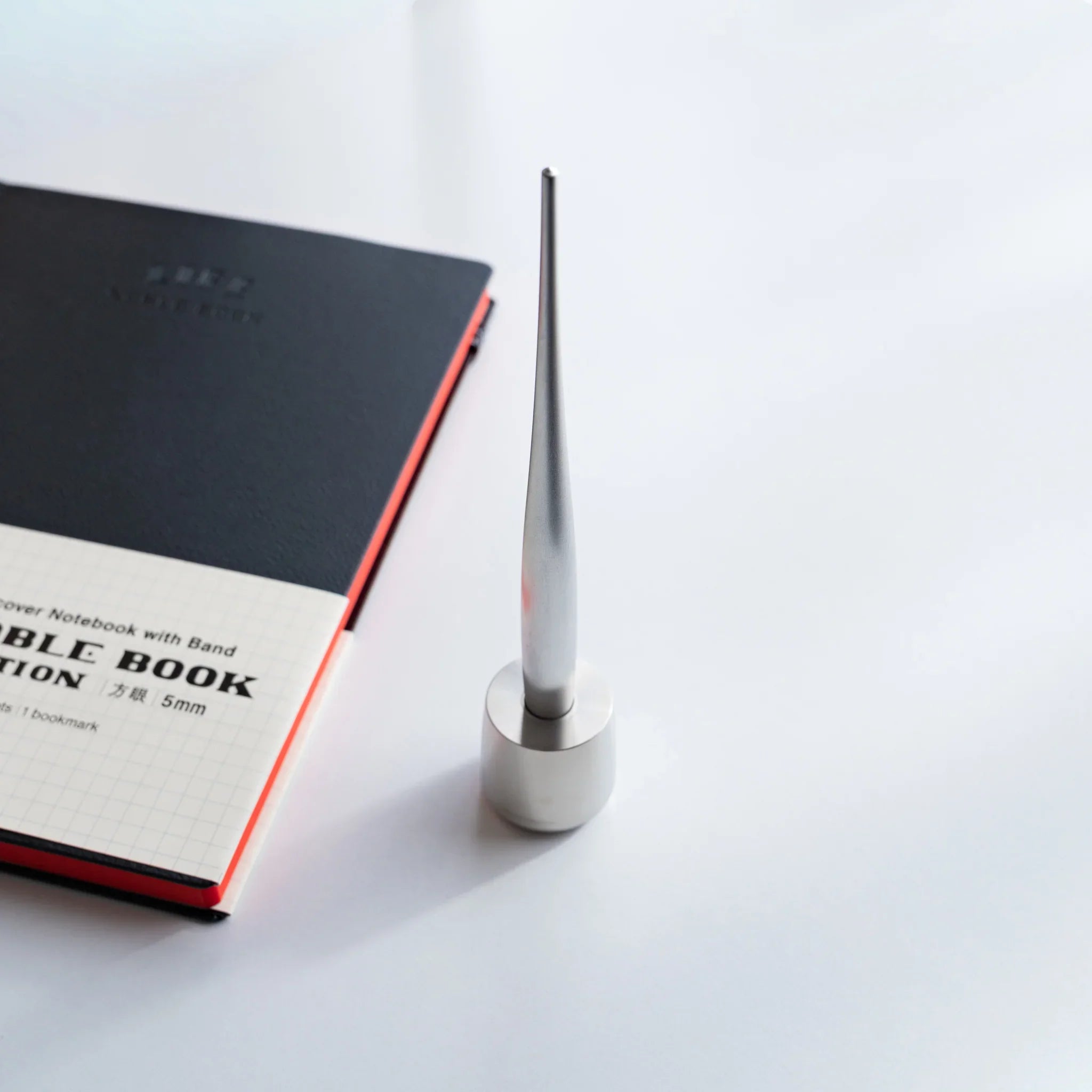 Japanese ballpoint pen with sleek brushed metal finish resting in stainless steel pen holder beside grid notebook