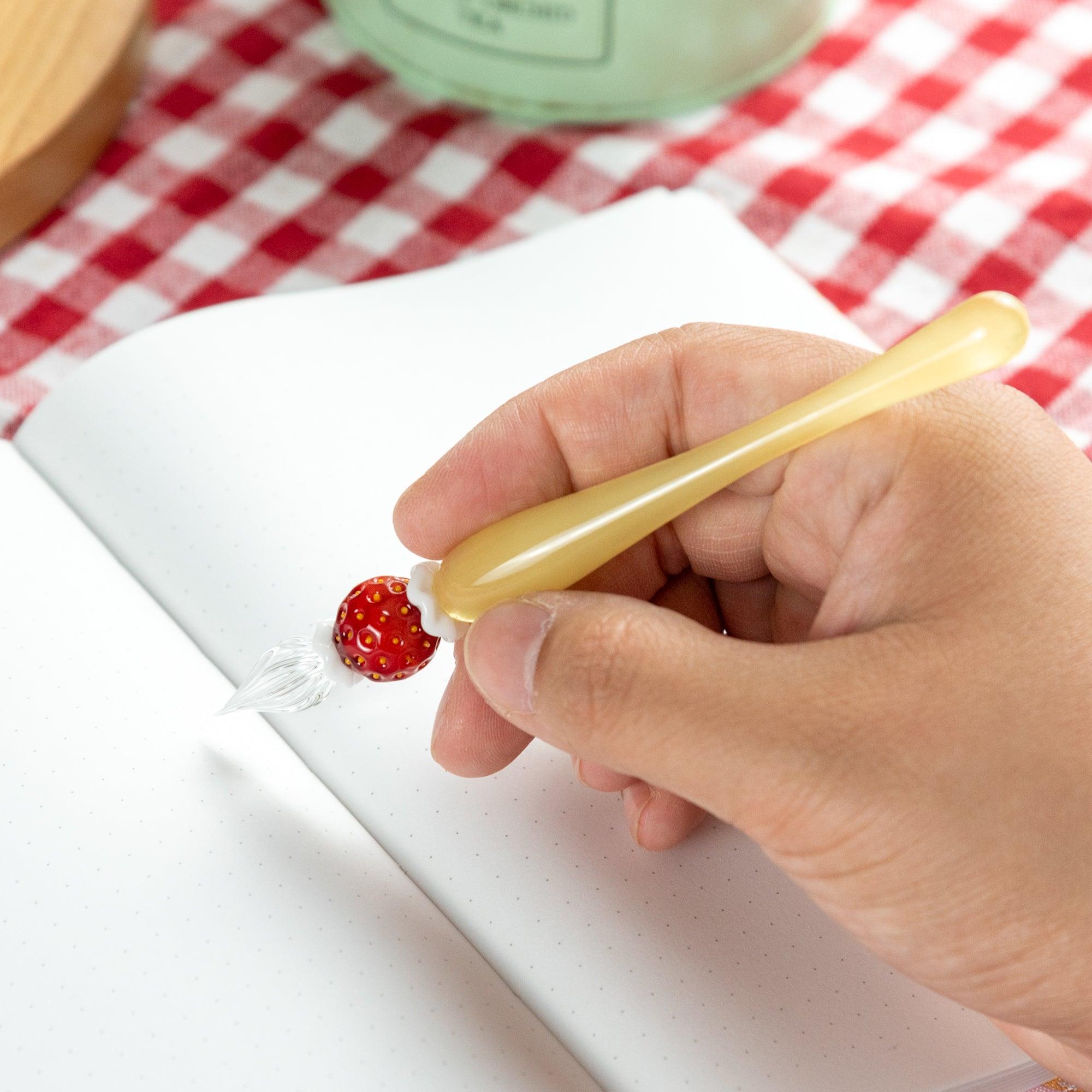 Hand holding a glass pen with a strawberry motif, writing on an open notebook. Perfect for Japanese stationery fans and collectors.