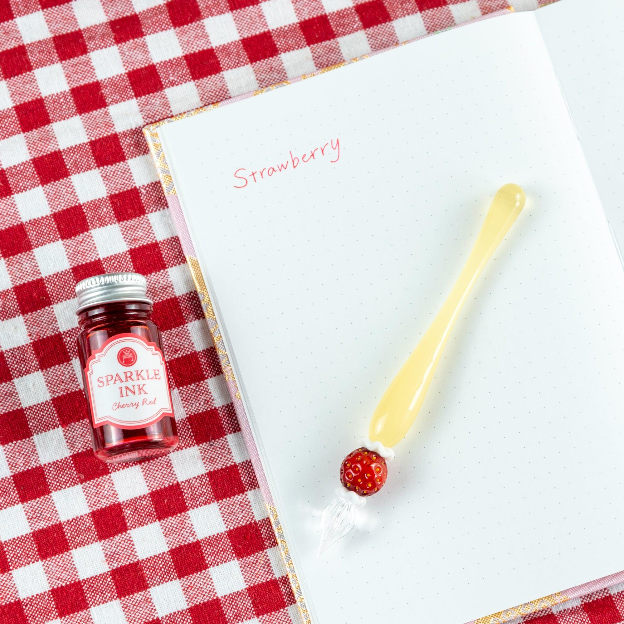 Glass pen with a strawberry motif and a bottle of red sparkle ink, placed on an open notebook. Great for stationery lovers.