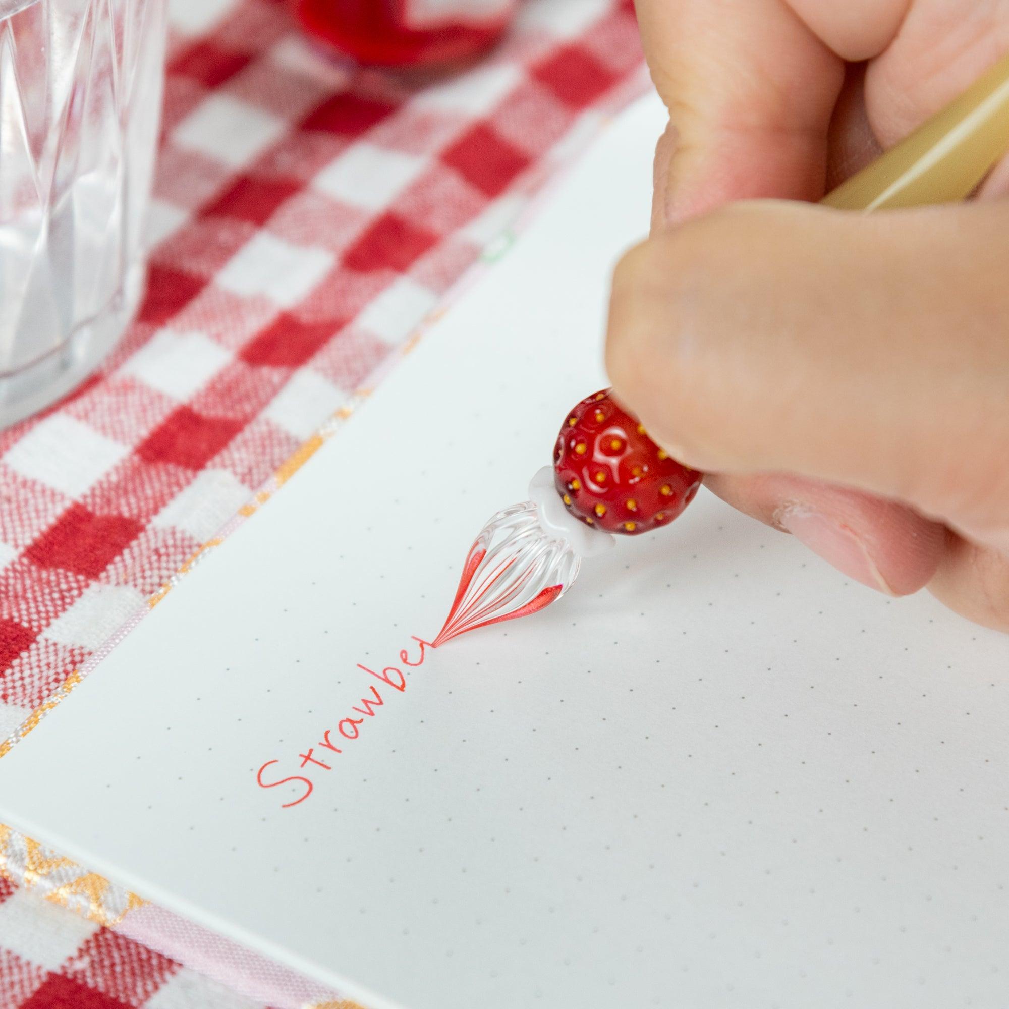 Strawberry-themed glass pen in action, writing red ink on a notebook. Ideal for Japanese stationery enthusiasts.