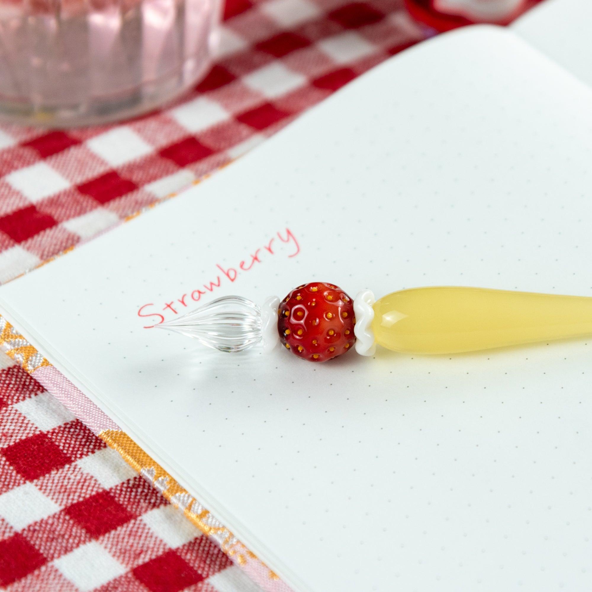 Strawberry-themed glass pen on a notebook with the word 'strawberry' written in red ink. Ideal for stationery collectors.