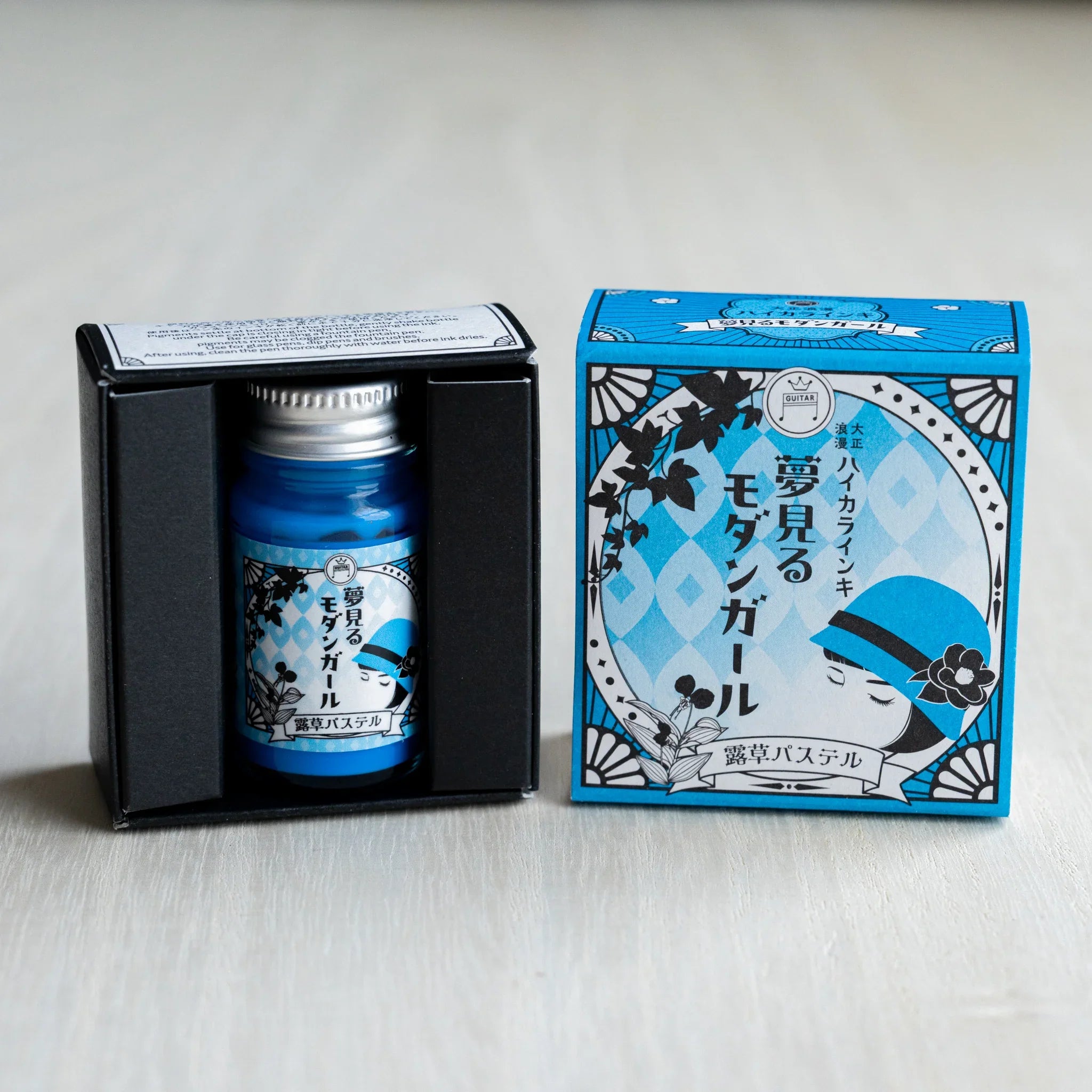 Taisho Romance Dayflower Pastel ink bottle in its open black inner box, next to the closed cyan blue outer box