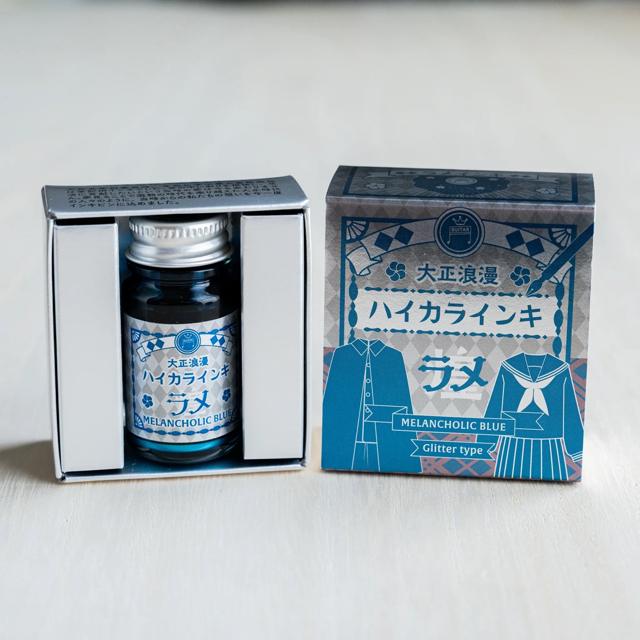 Taisho Romance Melancholic Blue Shimmer Ink bottle in its open black inner box beside the closed silver foil outer box
