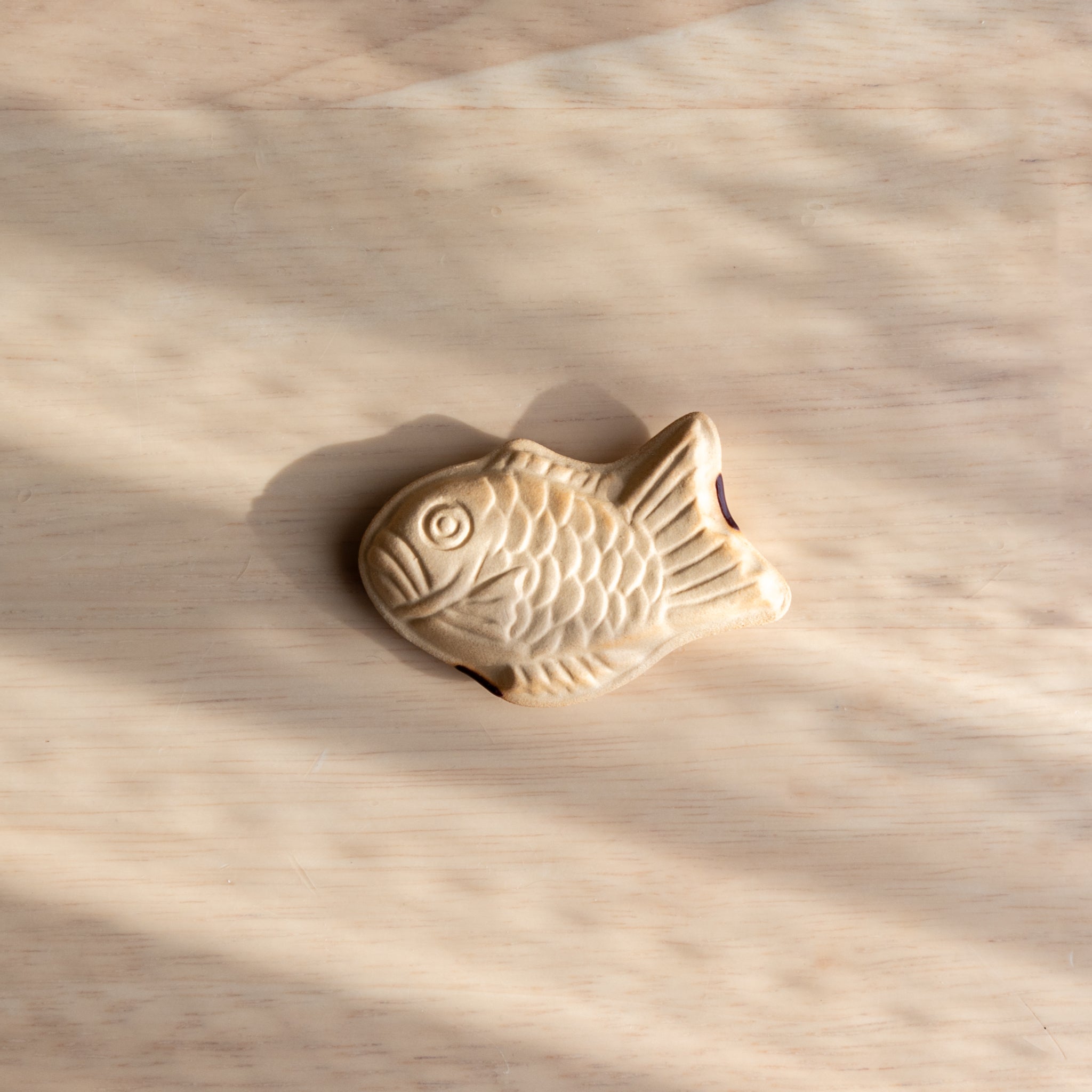 A fish-shaped Taiyaki Japanese ceramic pen rest in beige, placed on a wooden surface under soft sunlight.