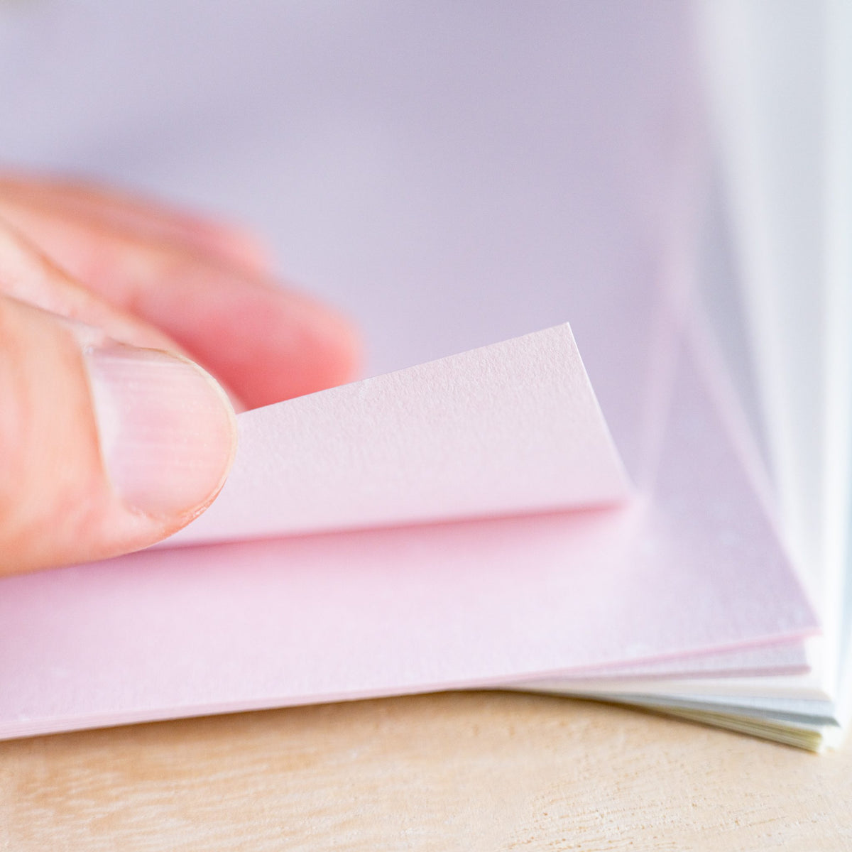 Fingertip lifting the corner of a pale pink Feather Waltz sheet showing fine fiber flecks in the paper