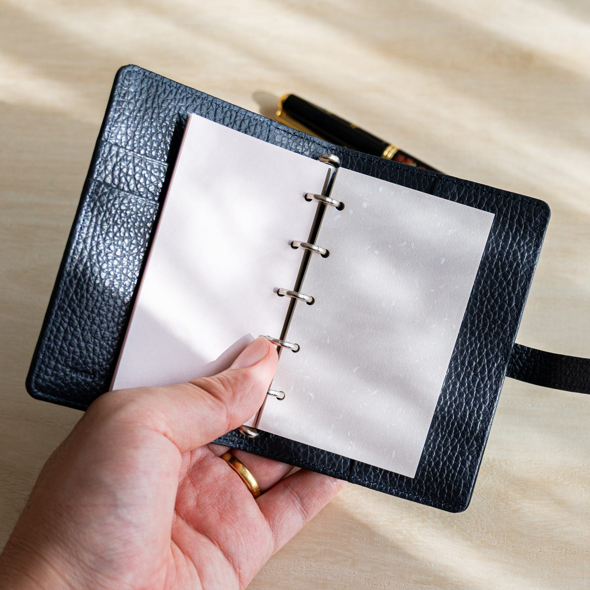 Hand holding an open navy planner with pale pink Feather Waltz M5 refill pages, fountain pen behind