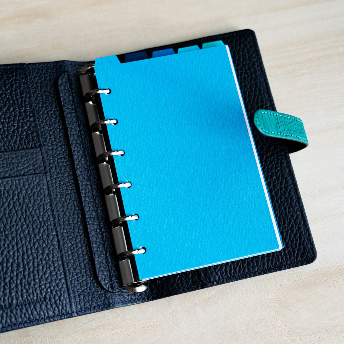 Sky blue Mermaid Mini 6 divider on the silver rings of an open navy planner with a teal snap tab