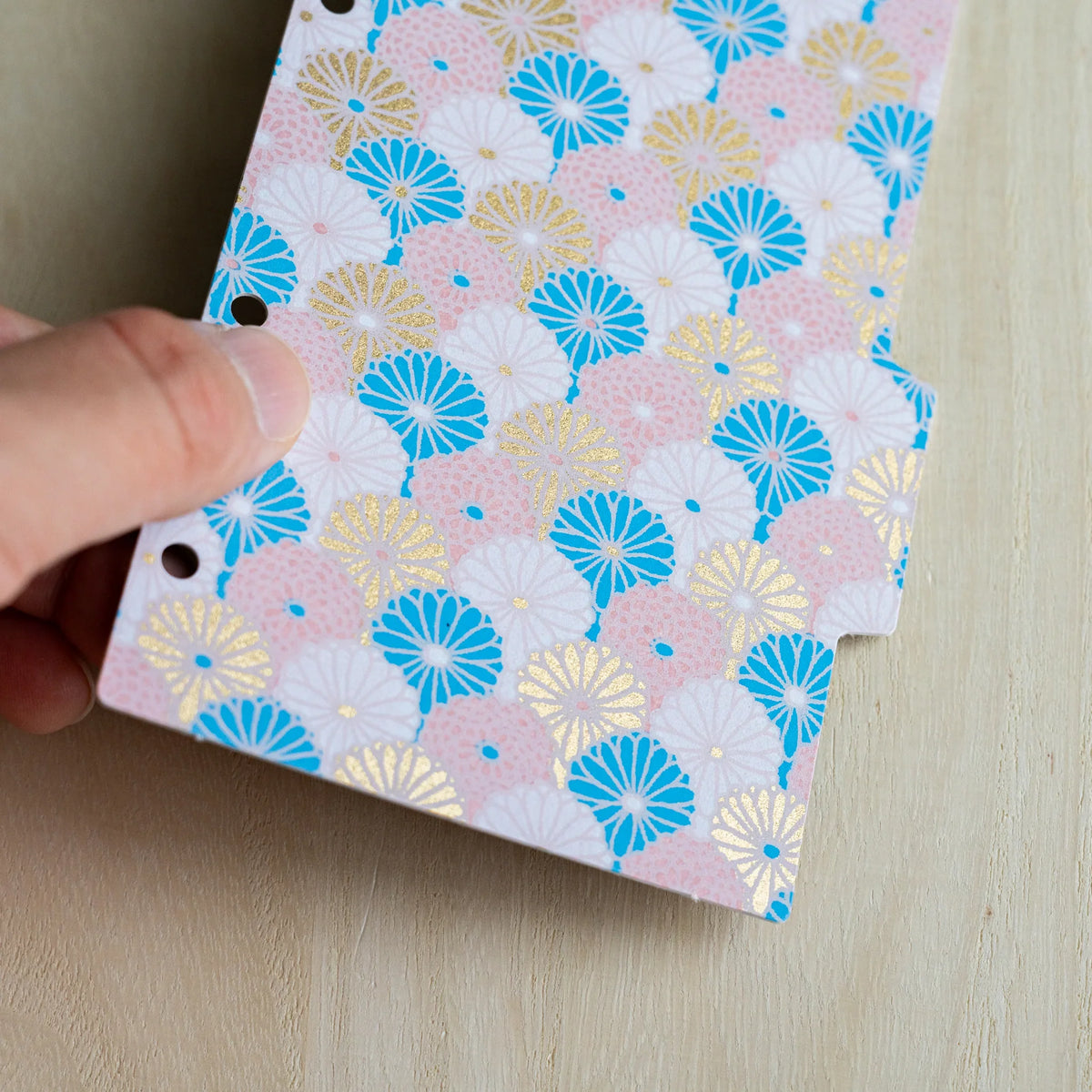 Hand holding a Tone index divider, close-up of pink, blue, white and gold chrysanthemum fans on washi