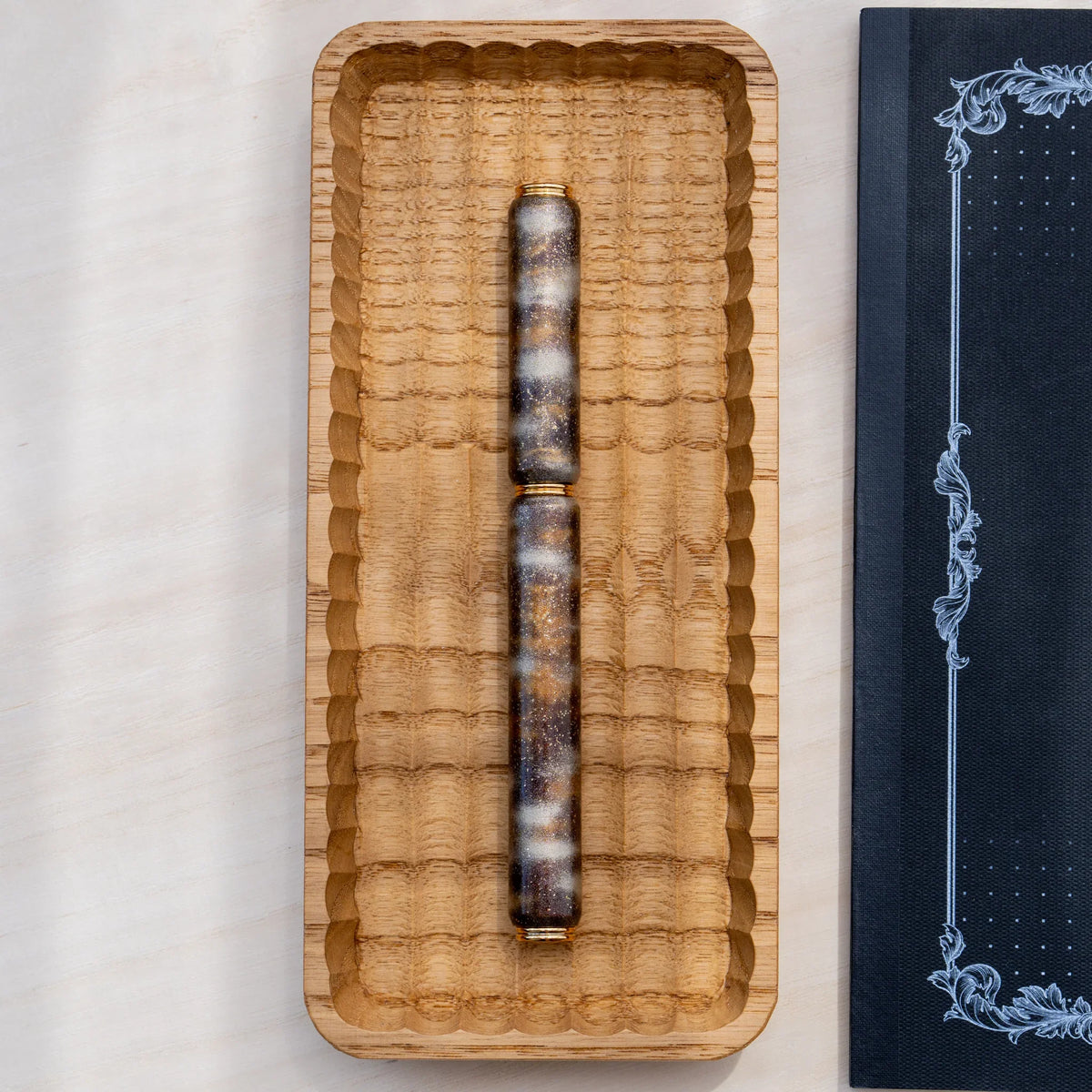 Translucent brown marbled urushi lacquered rollerball pen in zelkova wood, shown in a carved wooden tray with a Japanese notebook