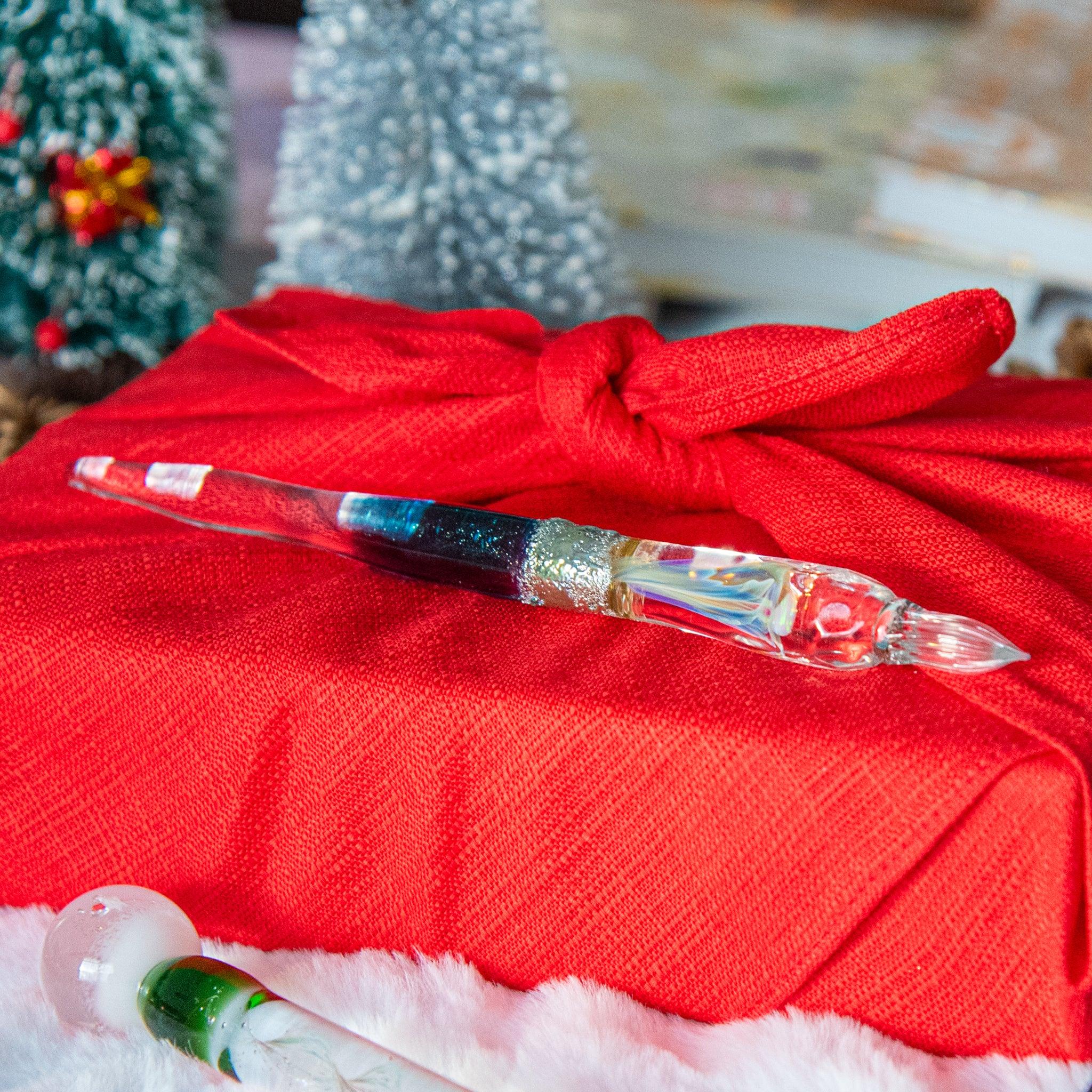 A glass pen featuring vibrant blue and silver accents rests on a red cloth-wrapped gift, with Christmas decorations in the background.