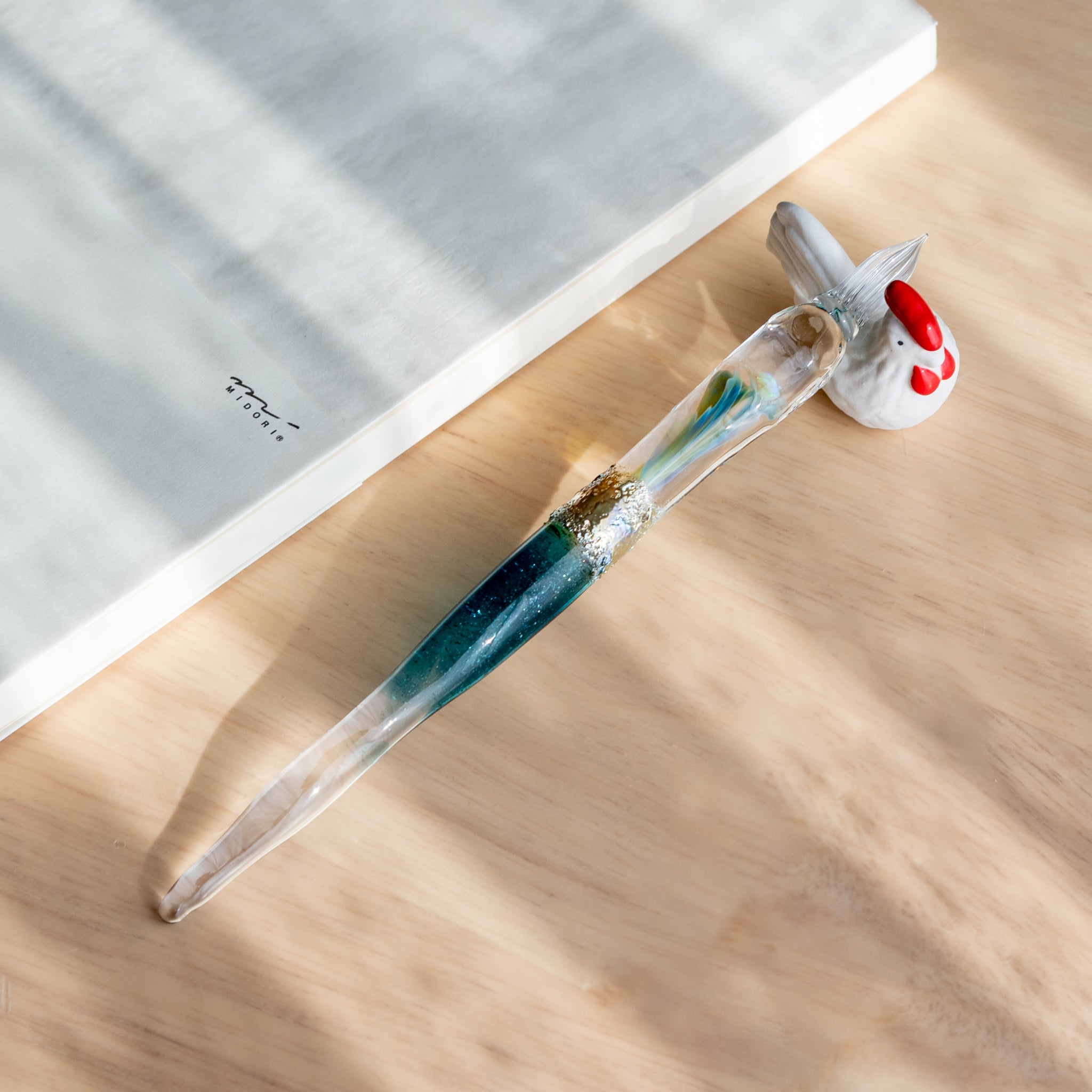 A Japanese glass dip pen with embedded opal and shimmering blue-green hues rests on a bird-shaped ceramic holder beside a white MD notebook on a wooden desk.  