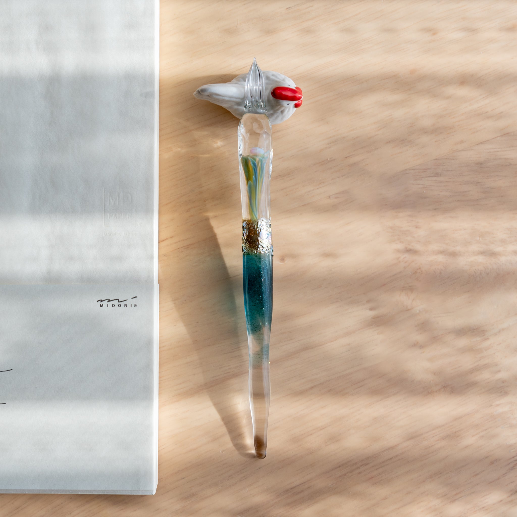 A Japanese glass dip pen with embedded opal and swirling colors rests on a bird-shaped ceramic holder. A white MD notebook lies nearby on a wooden desk. 