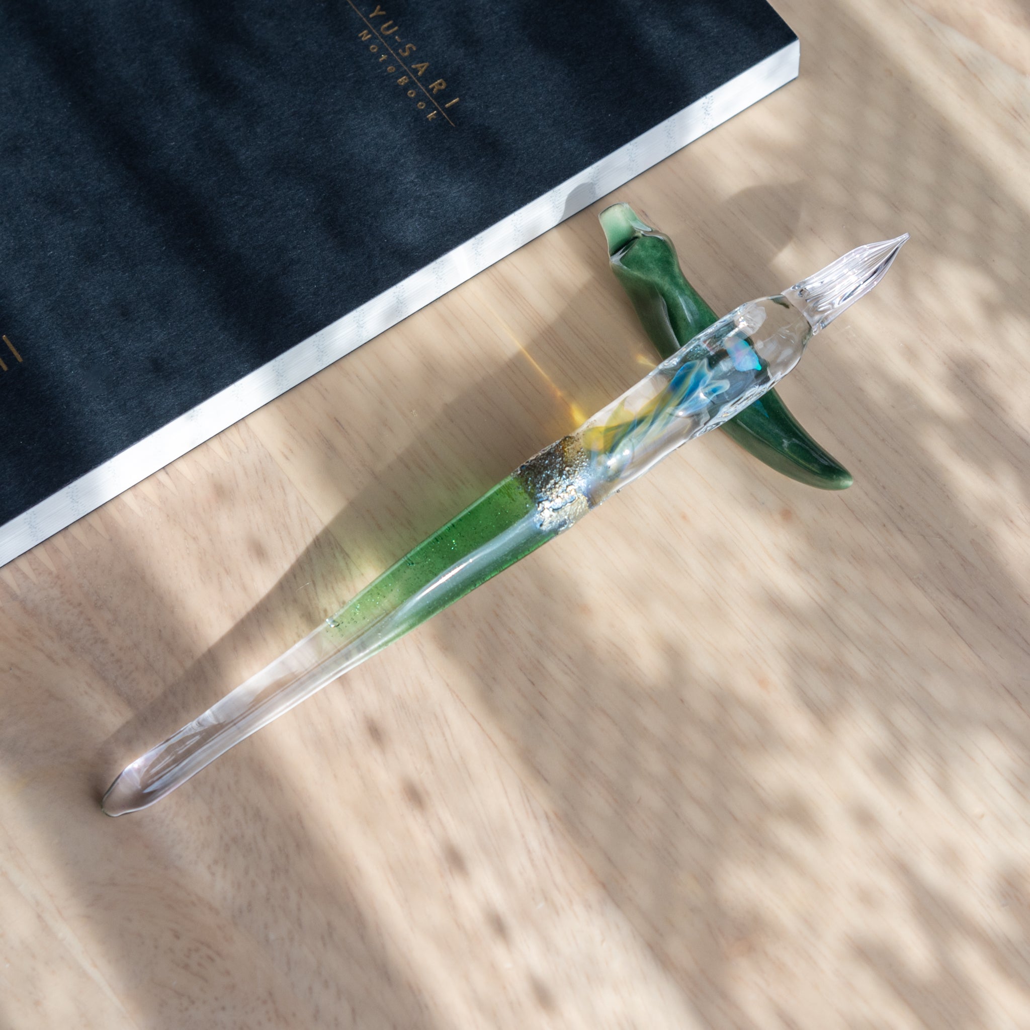 A Japanese glass dip pen with embedded opal and green gradient rests on a green chili-shaped ceramic holder. A black Yusari notebook lies nearby on a wooden desk.