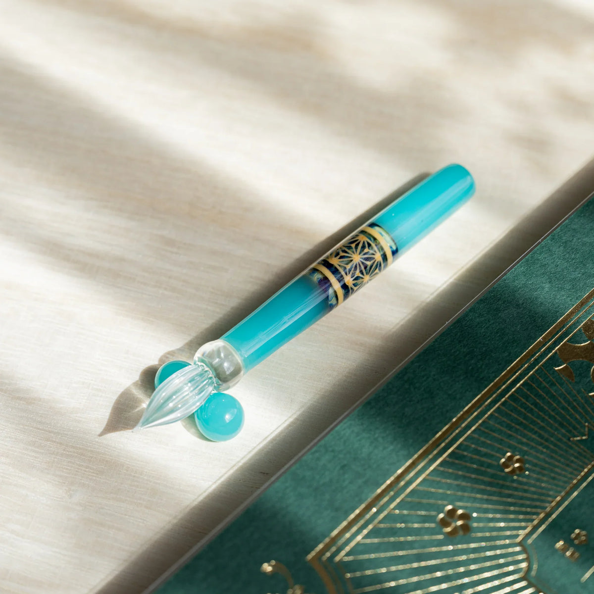 Turquoise Hemp Leaves glass dip pen angled across pale wood, hemp leaf band visible on the barrel