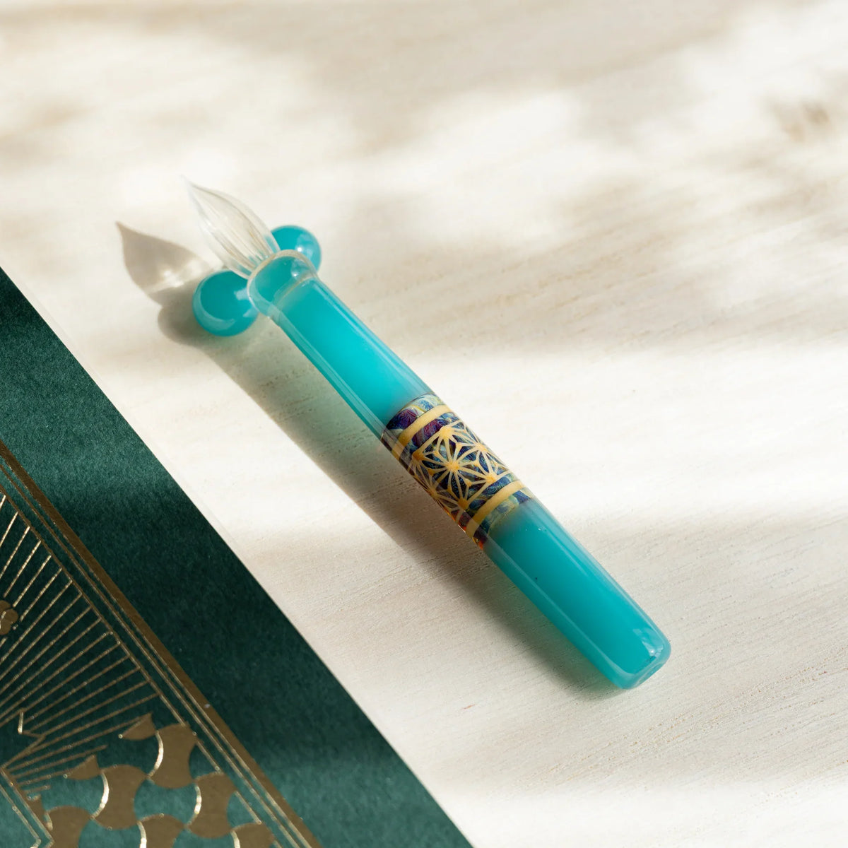 Turquoise Hemp Leaves glass dip pen angled on pale wood, nib resting on a turquoise glass sphere