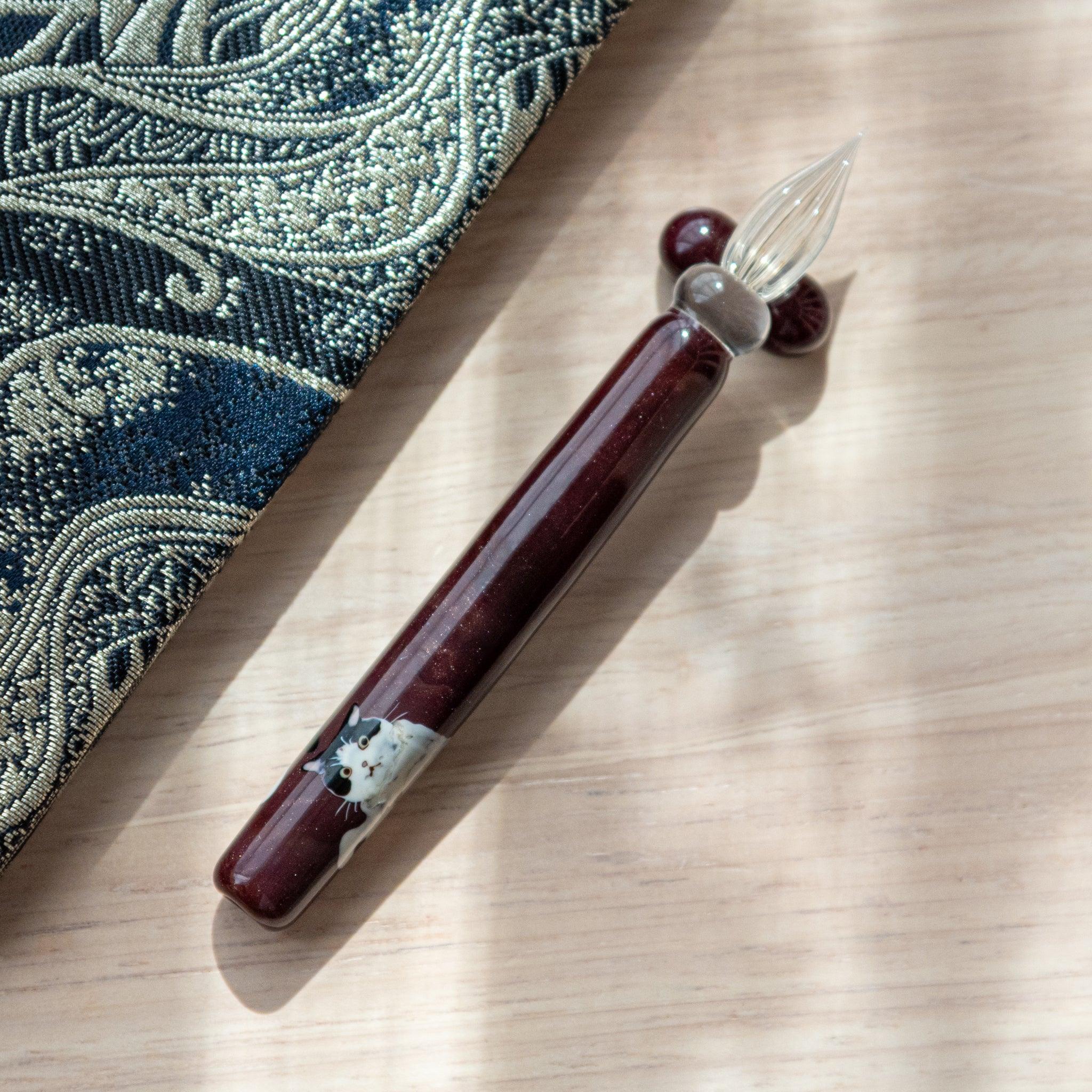 Dark red glass pen featuring a Tuxedo cat design, paired with a matching glass pen rest. Displayed on a wooden surface beside a Nishijin textile notebook.