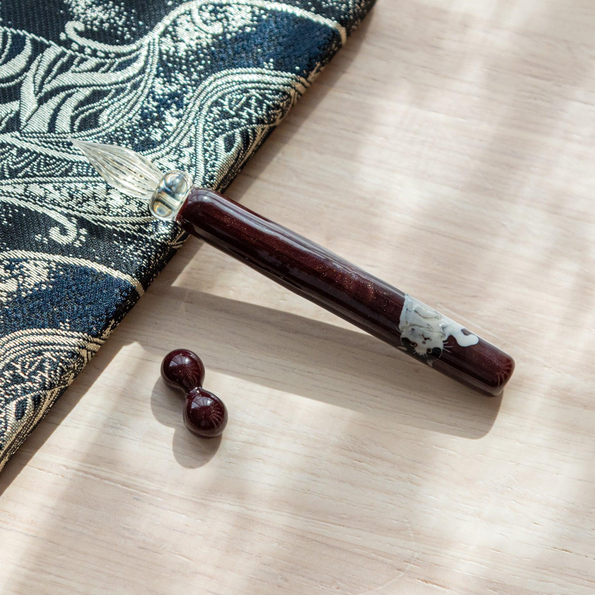 Dark red glass pen featuring a Tuxedo cat design, paired with a matching glass pen rest. Displayed on a wooden surface beside a Nishijin textile notebook.