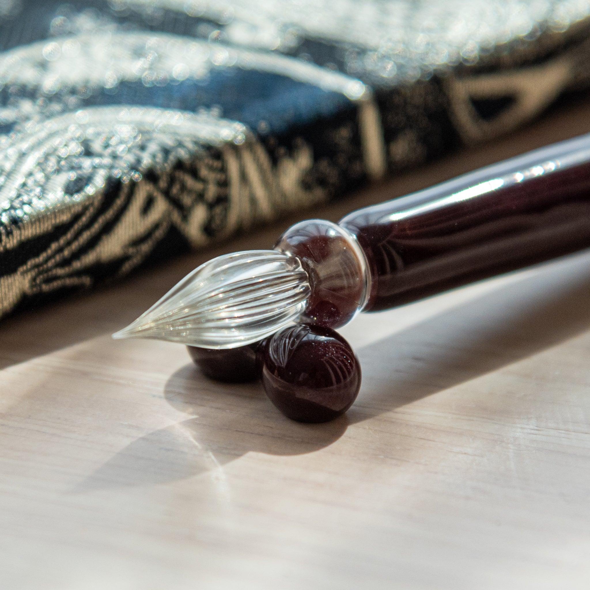 Dark red glass pen featuring a Tuxedo cat design, paired with a matching glass pen rest. Displayed on a wooden surface beside a Nishijin textile notebook.