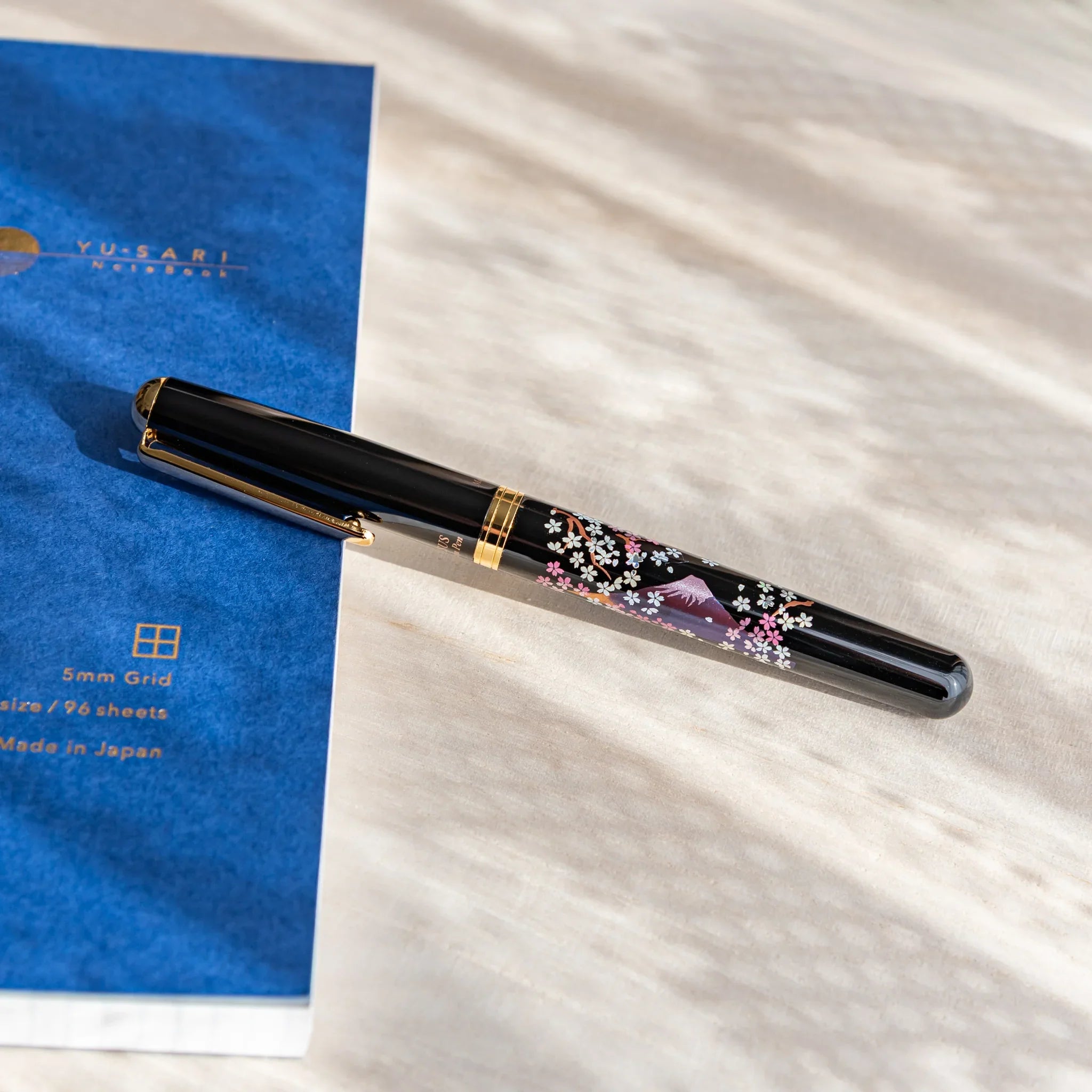 Black Japanese fountain pen with pink sakura and Mt. Fuji artwork resting on a light desk beside a blue notebook.