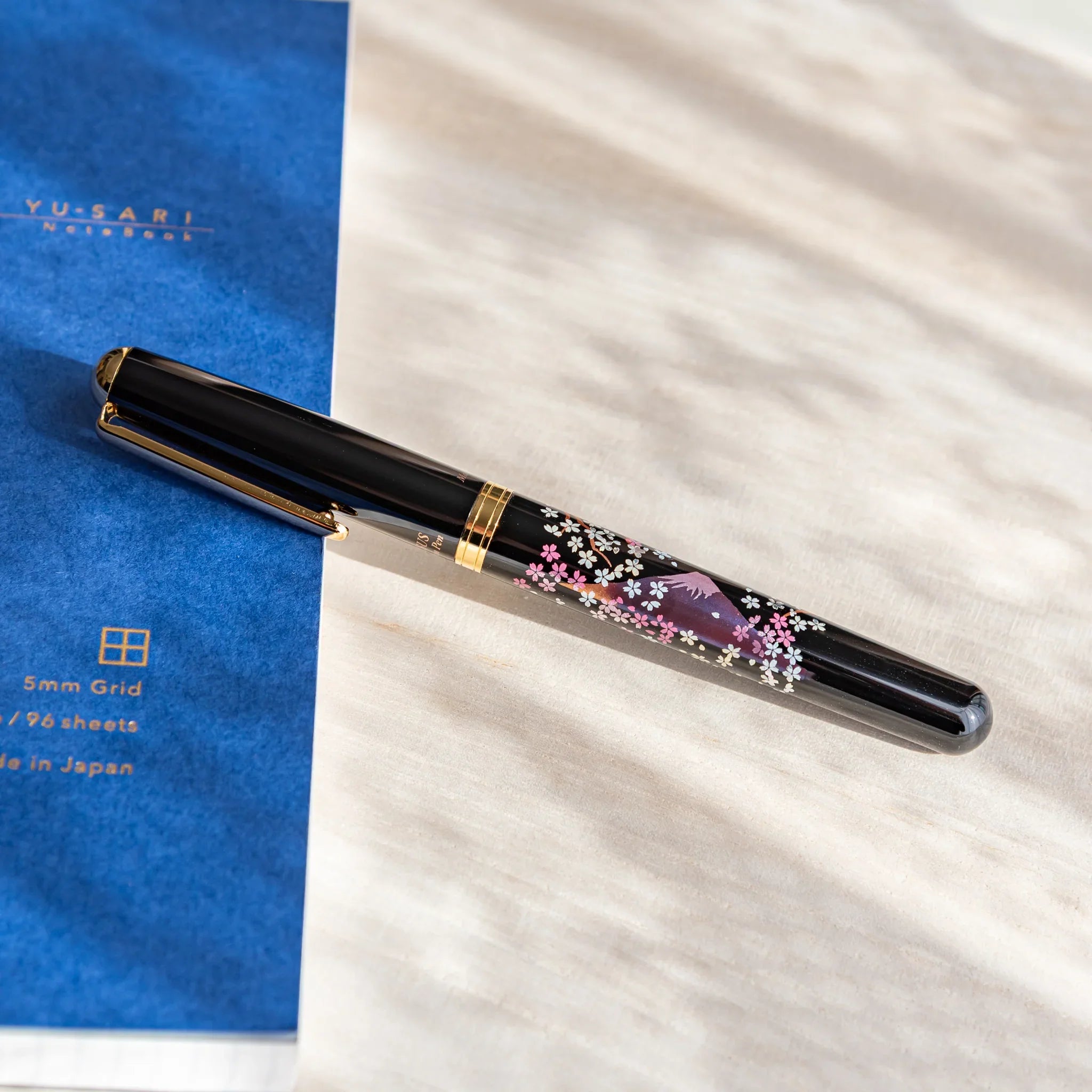 Black Japanese fountain pen with pink sakura and twilight Fuji artwork resting on a light desk beside a blue notebook.