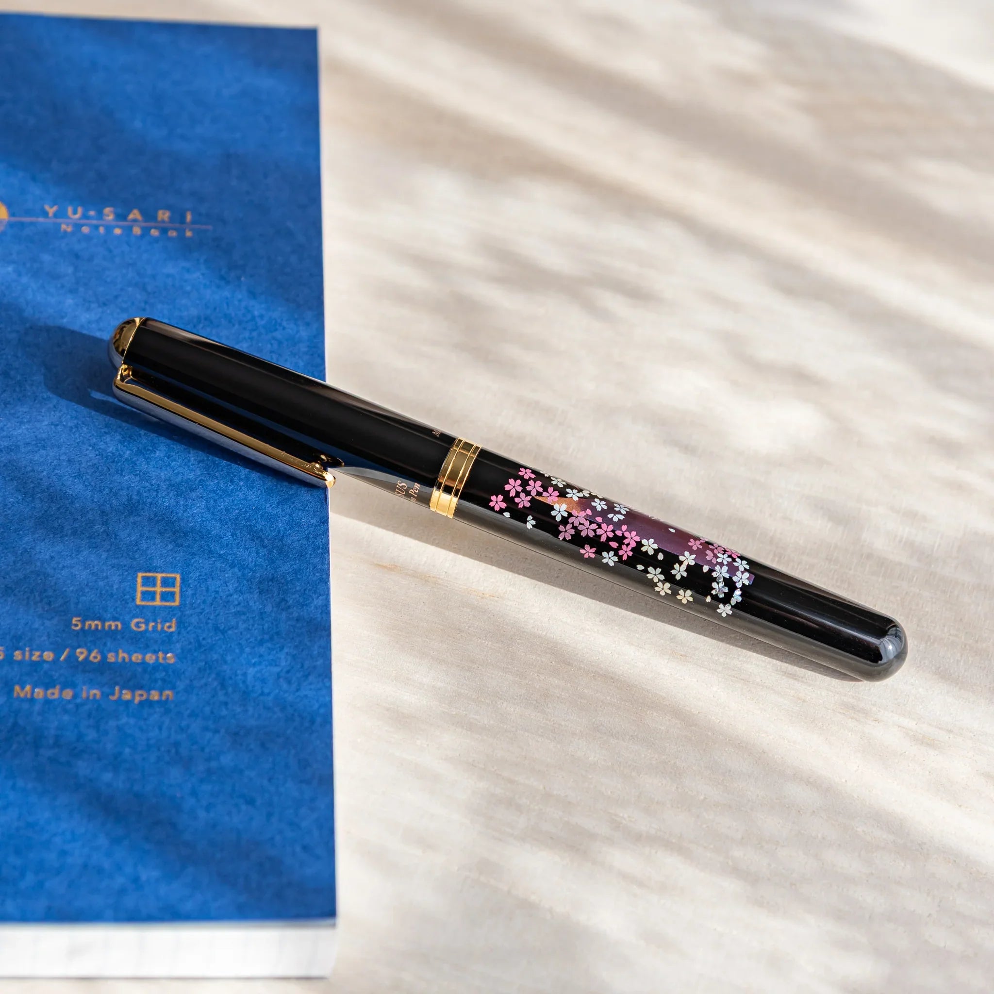 Black Japanese fountain pen with pink sakura and twilight Fuji artwork resting beside a blue notebook.