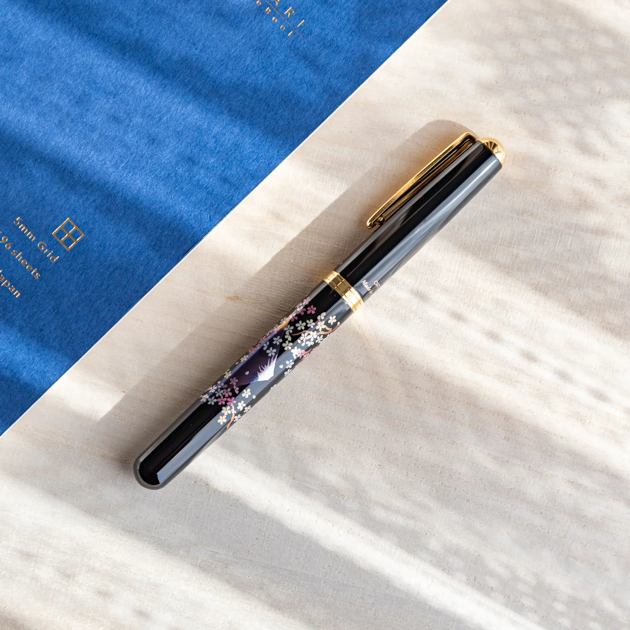 Japanese fountain pen with sakura and twilight Mt. Fuji artwork resting on a light desk beside a blue notebook.