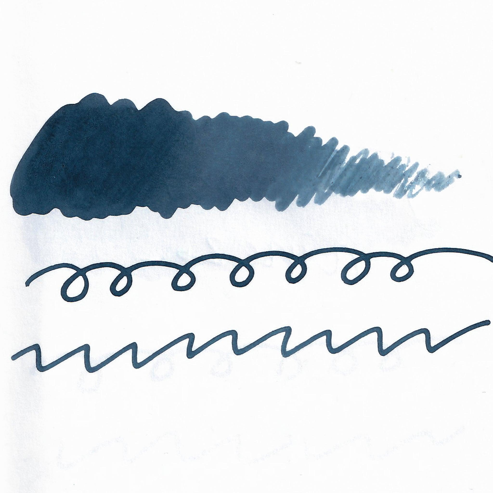 Swatch of blue-gray fountain pen ink on white paper. The ink shows shading variations, with a dark center and lighter edges. Below are wavy and looped lines demonstrating the ink's flow.  