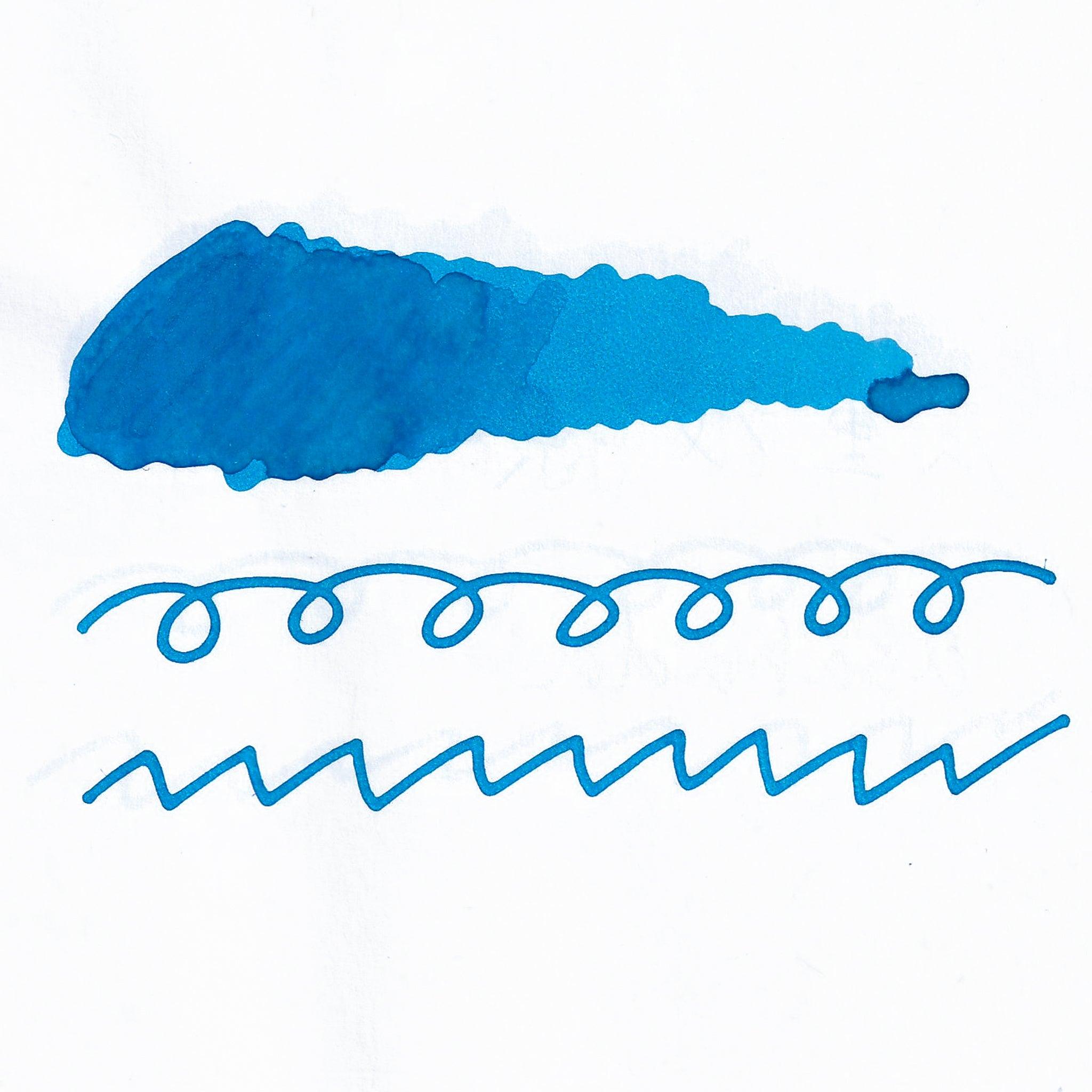 Swatch of light blue fountain pen ink on white paper. The ink shows shading variations, with a dark center and lighter edges. Below are wavy and looped lines demonstrating the ink's flow.  