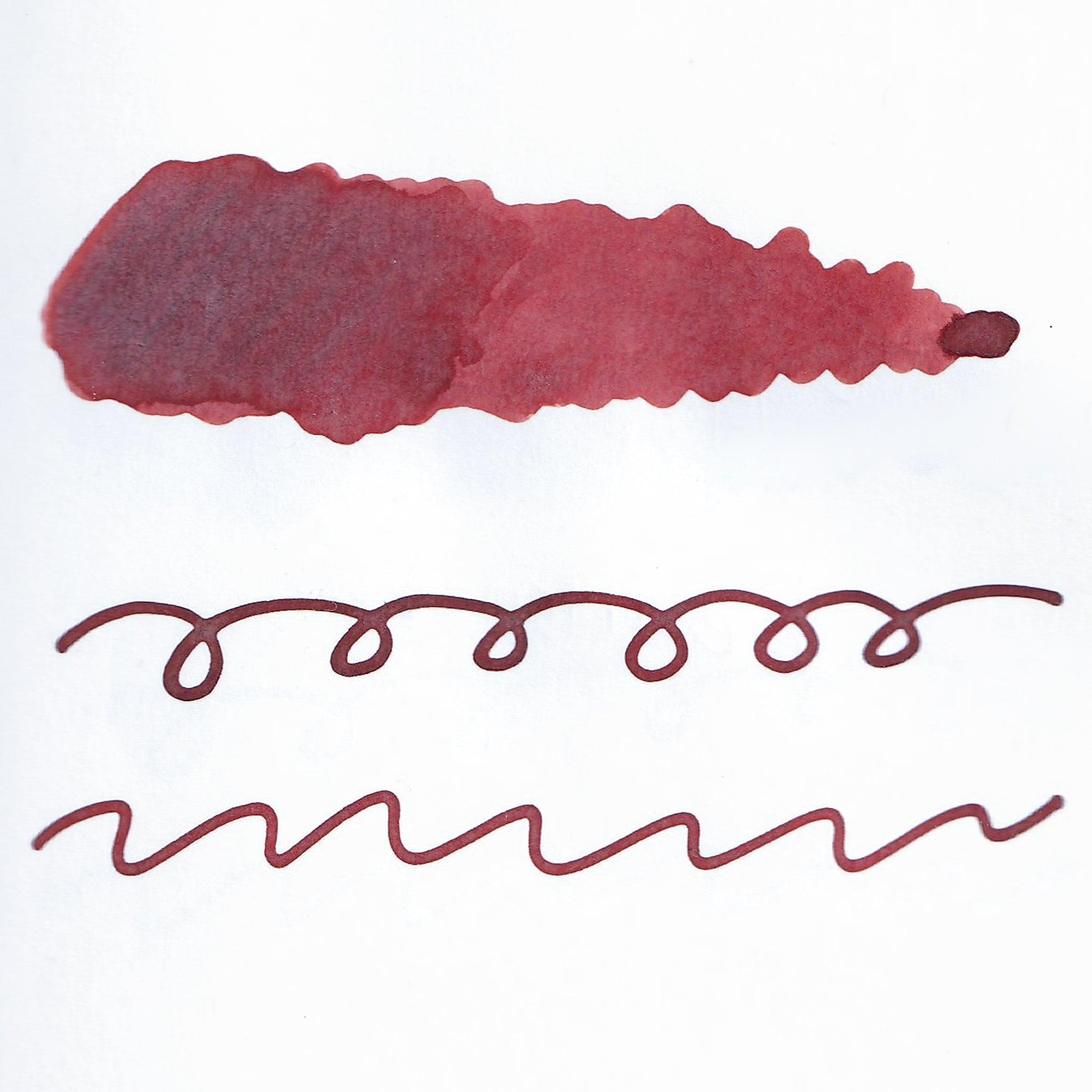 Swatch of red fountain pen ink on white paper. The ink shows shading variations, with a dark center and lighter edges. Below are wavy and looped lines demonstrating the ink's flow.  