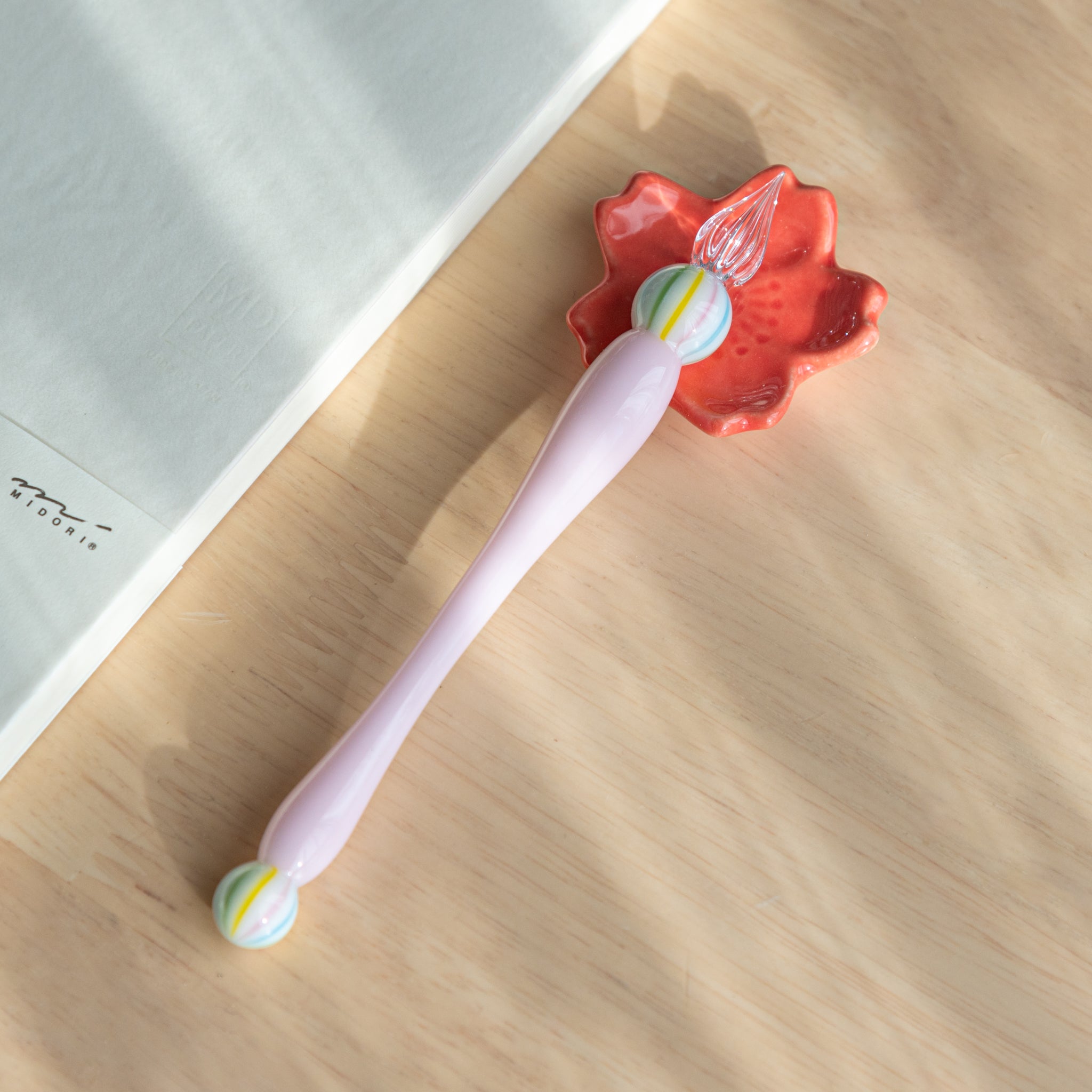 A glass dip pen, a Japanese stationery invention, with a pink handle and glass marble, rests on a red maple leaf-shaped holder on a wooden desk.  