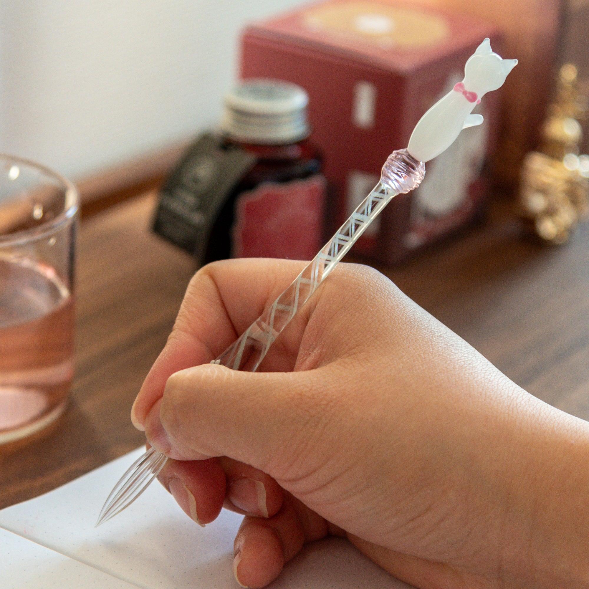 Hand holding a glass pen with a white cat design, writing in a notebook, showcasing the pen's smooth and elegant use.