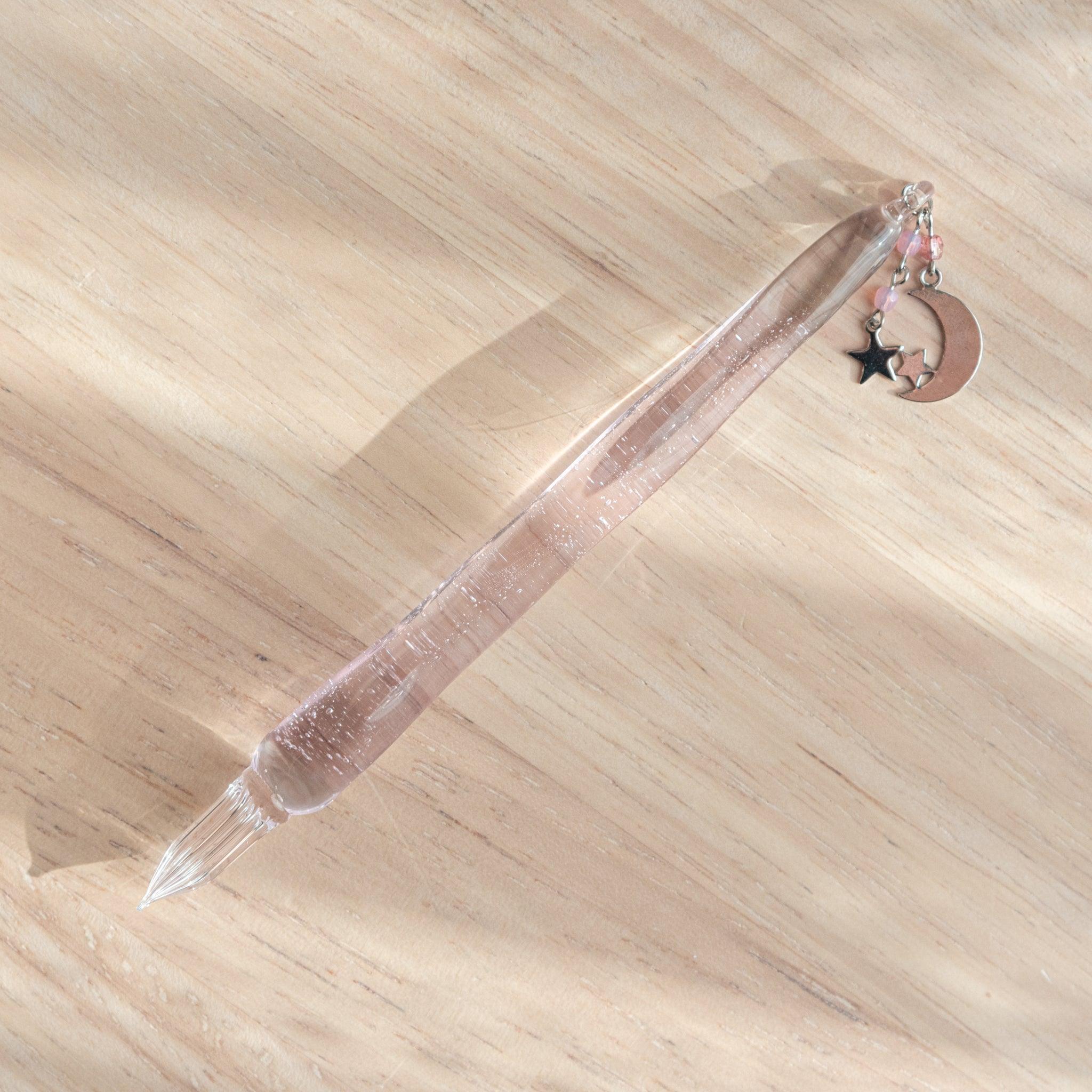 Transparent glass dip pen with subtle pink hue, accented with moon and star charms and pink beads, placed on a light wooden surface.