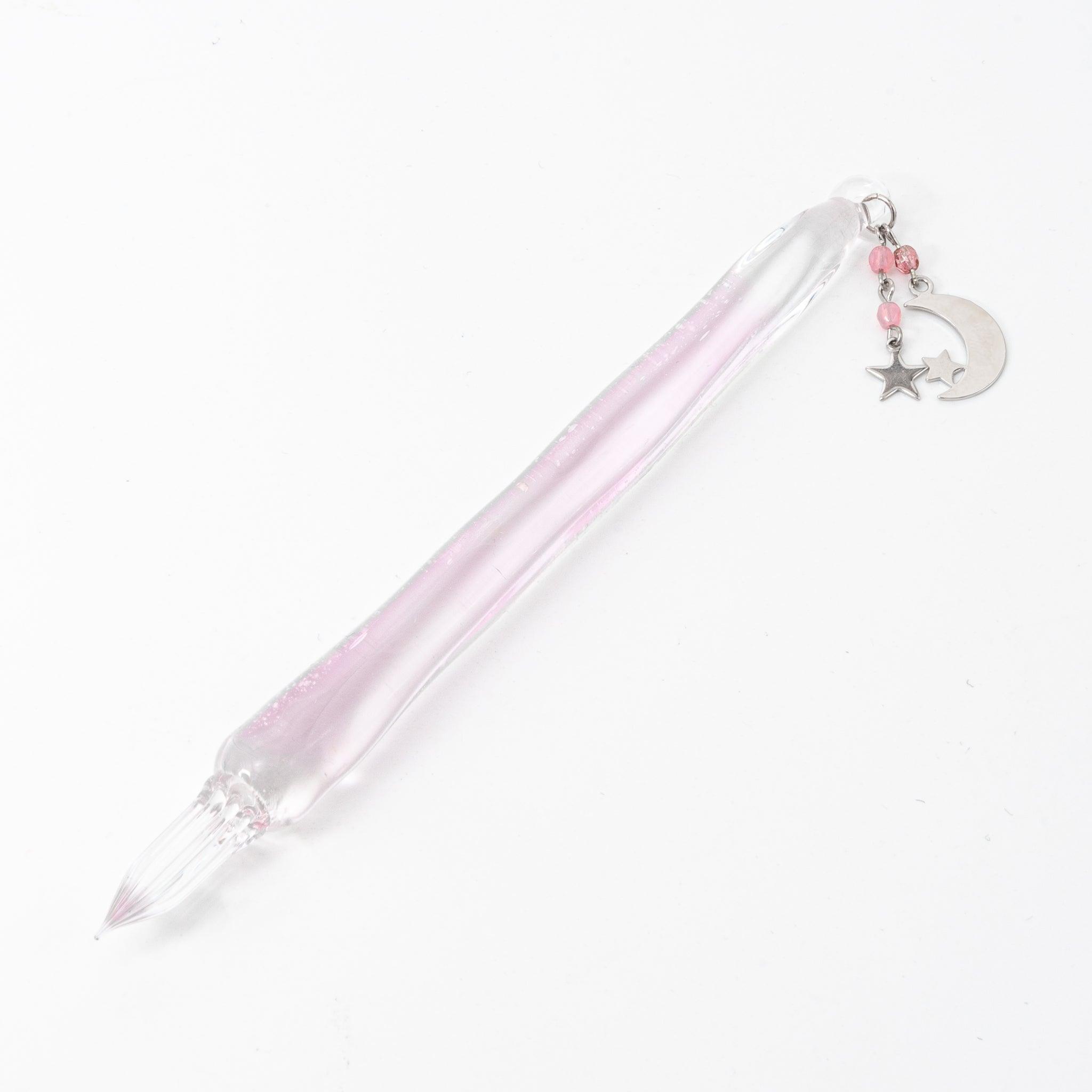 Clear glass dip pen with a faint pink tint, featuring crescent moon and star charms adorned with small pink beads, lying on a white background.